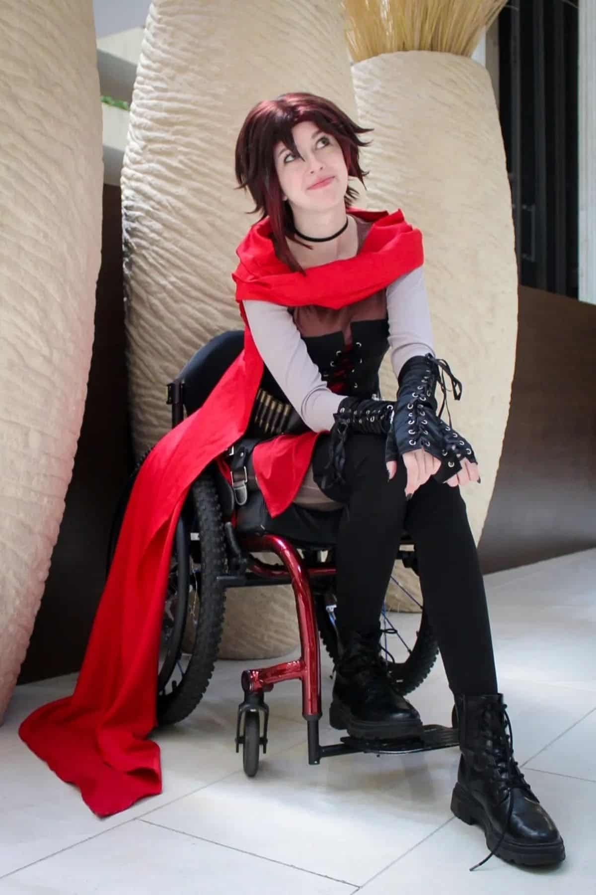 Person in a red cape cosplay sits in a wheelchair indoors, wearing black boots and gloves, with neutral expression.
