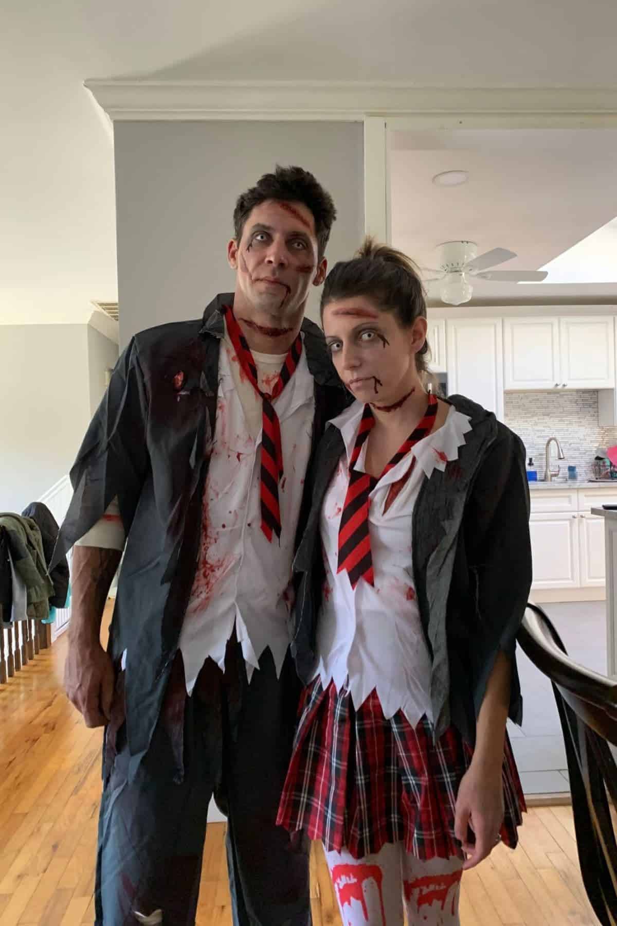Two people in matching zombie school uniforms with fake blood and makeup stand together in a bright kitchen.
