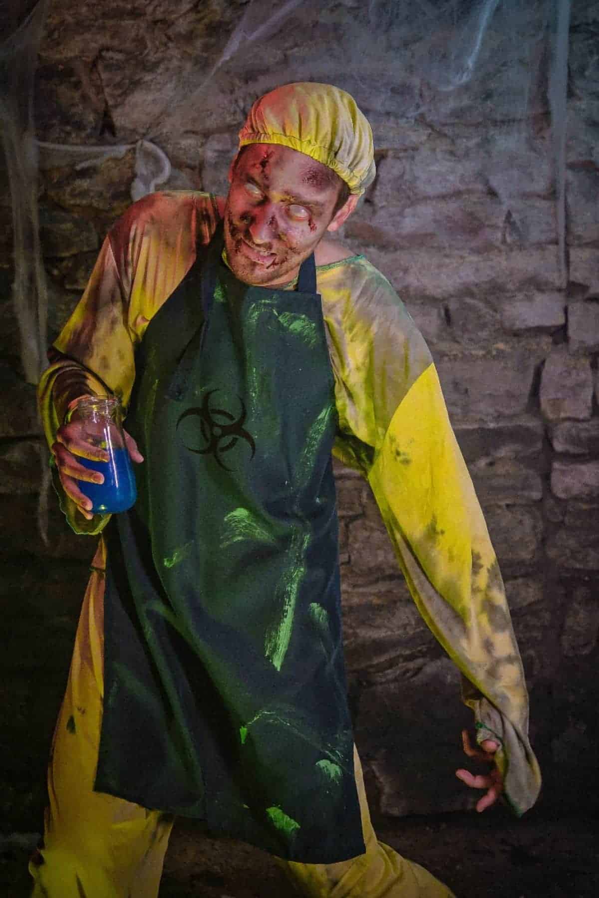 Person in a yellow hazmat suit and dark apron with a biohazard symbol, holding a jar of blue liquid, appears to be acting as a zombie in a dimly lit, stone-walled setting.