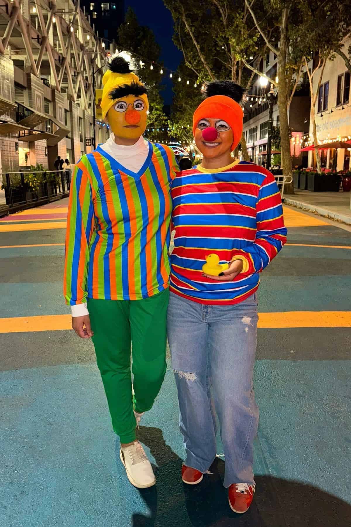 Two people dressed as Bert and Ernie from Sesame Street stand together on a colorful street, wearing striped shirts, beanies, and holding a rubber duck.