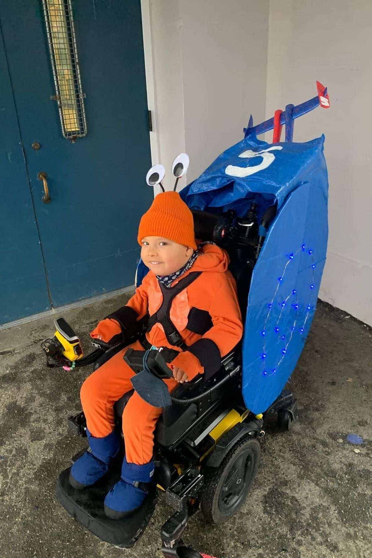 A child in an orange costume and hat with googly eyes sits in a wheelchair decorated as a blue snail with lights and a number 5 on its shell.