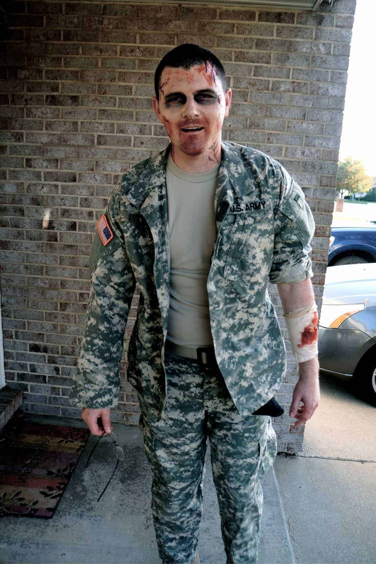 Person in a U.S. Army uniform with makeup and fake blood, appearing as a zombie, standing outside near a brick wall and parked car.