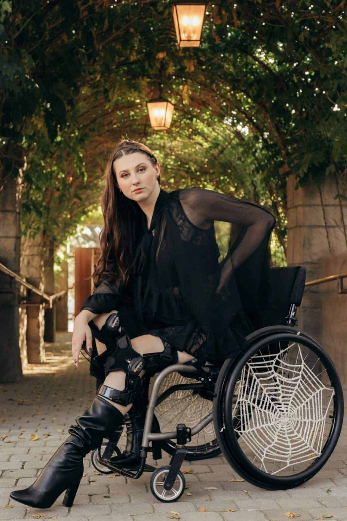 Woman in a black dress and boots sits in a wheelchair with spiderweb designs on the wheels, under an arched walkway with hanging lamps and greenery.