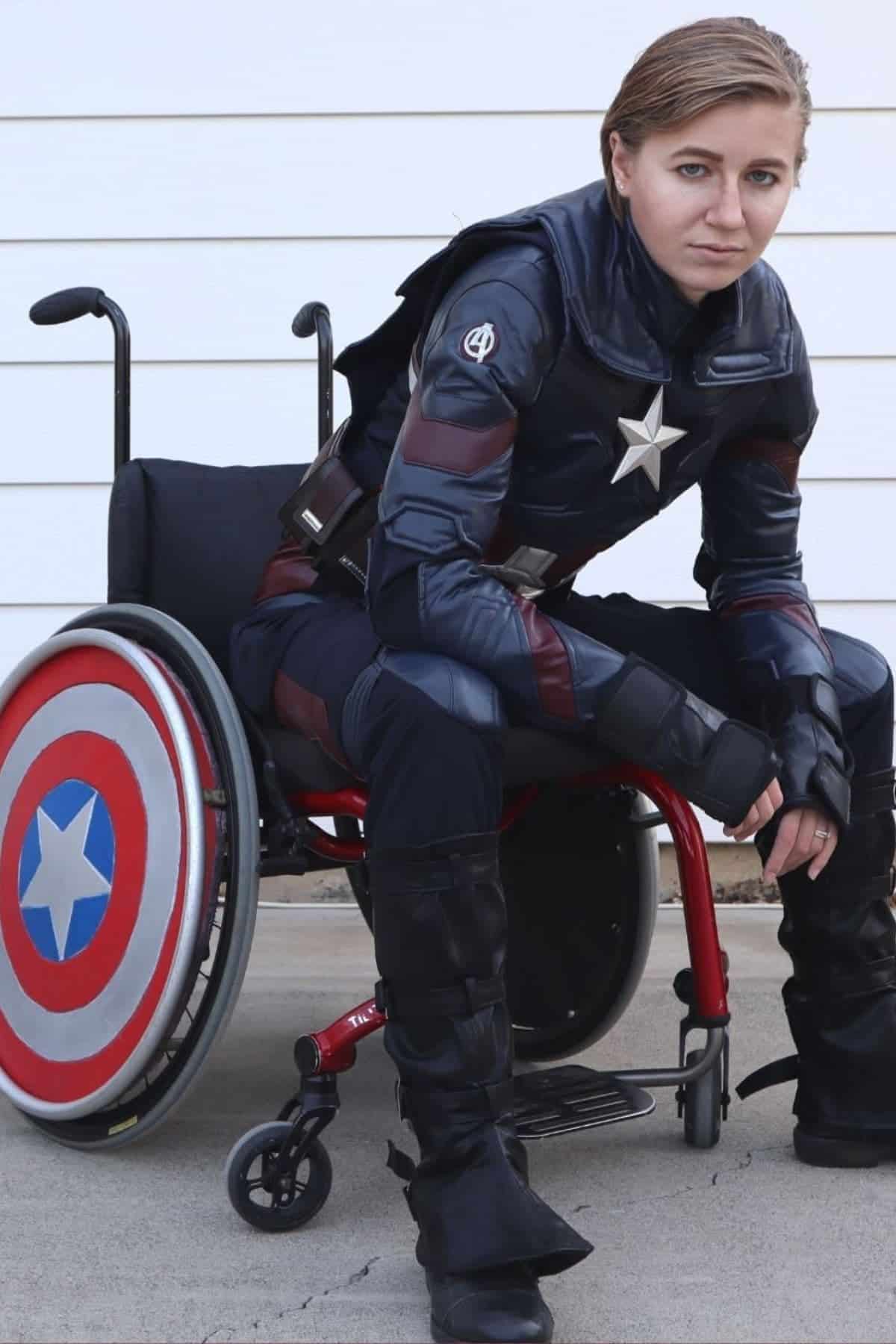 Person in a Captain America costume sits in a wheelchair, which has a Captain America shield attached to its wheel, against a white background.