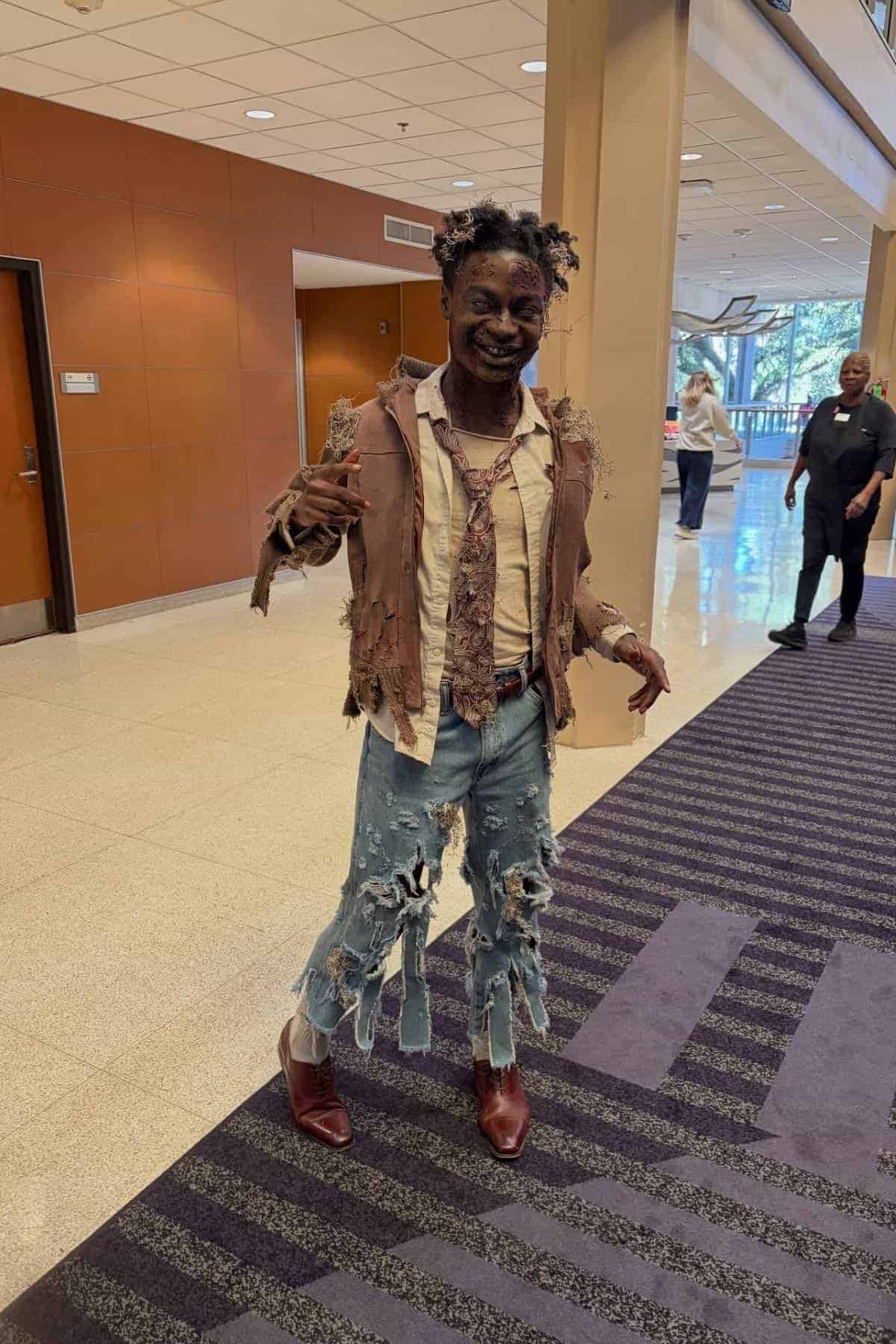 Person in ripped, tattered clothing and distressed jeans stands indoors, smiling and posing for the camera; background shows hallway and other people.
