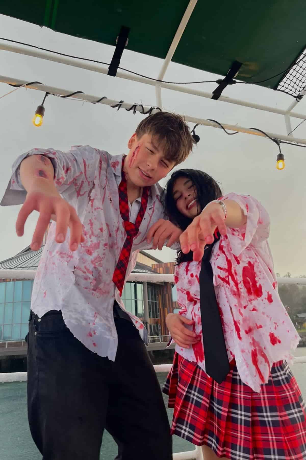 Two people in blood-splattered school uniforms pose with outstretched arms, imitating zombies, under string lights near a waterside building.