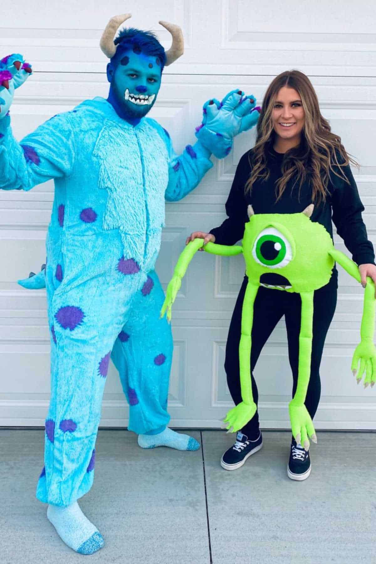 Two people in front of a garage; one in a blue monster costume with horns, the other in black clothes holding a green, one-eyed creature prop with long arms and legs.