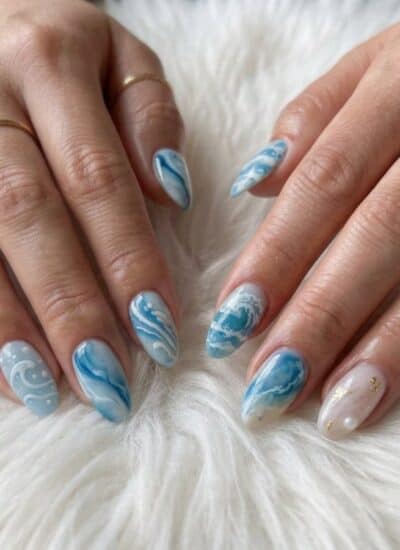 Two hands with almond-shaped nails featuring blue and white wave designs rest on a white fur surface. Three gold rings are worn on the fingers.