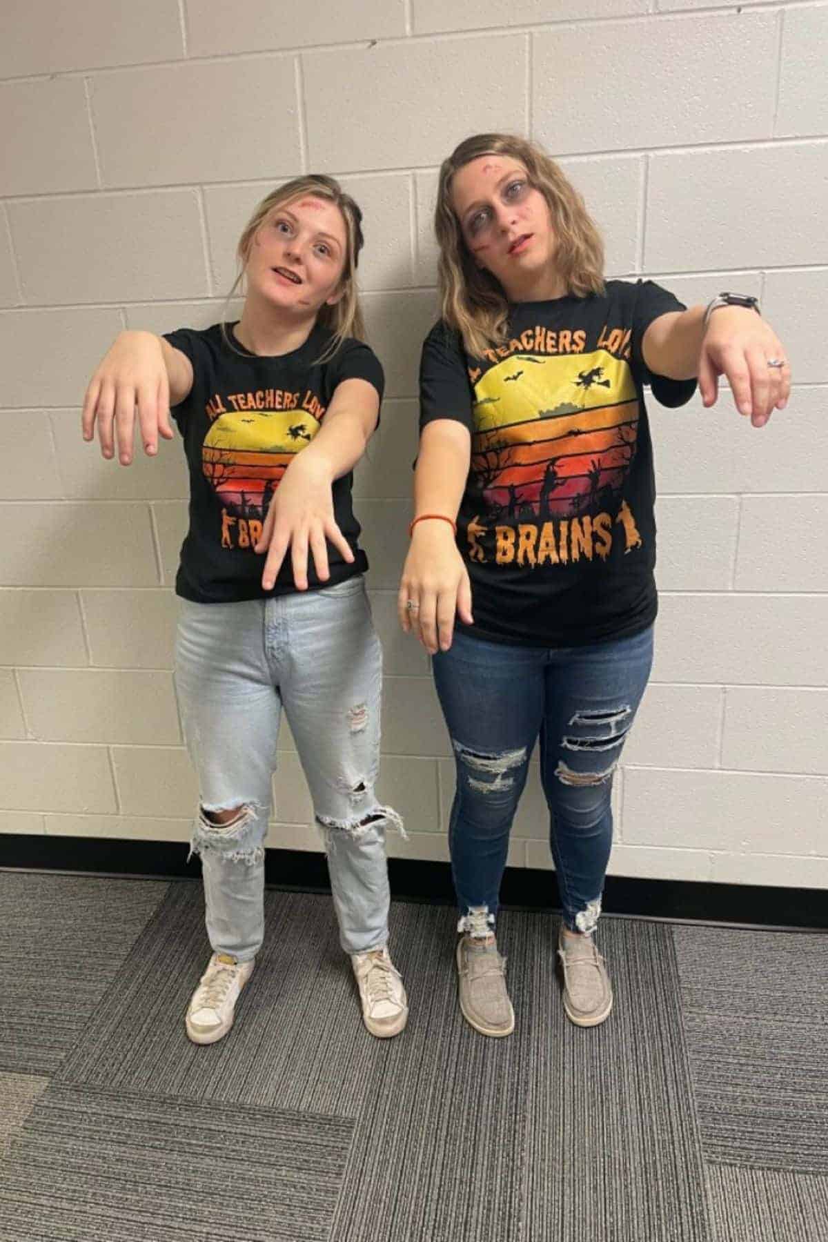 Two people dressed as zombies stand against a white brick wall, wearing matching “Teachers love brains” shirts and light jeans, with arms outstretched and faces painted.