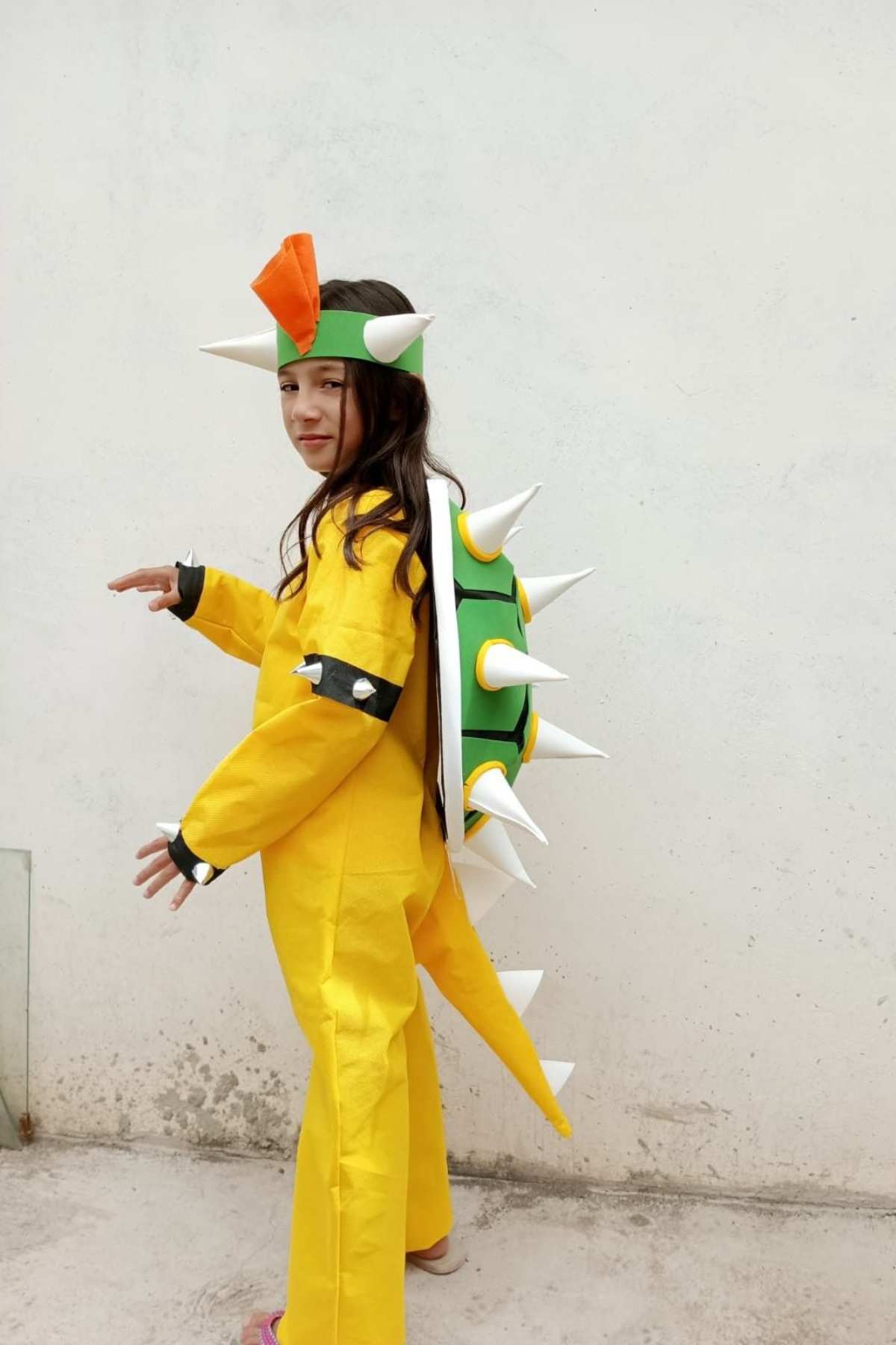 Child in a Bowser costume with spikes and a green shell, resembling Bowser from the Mario video game series, stands against a plain white wall.