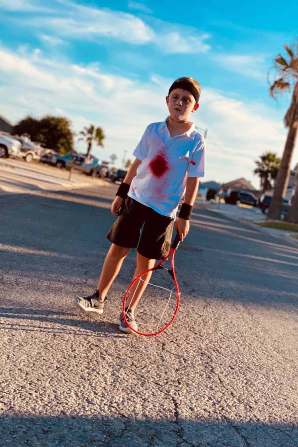 Boy in tennis attire stands on a street holding a broken racket, with a red stain resembling a fake injury on his white shirt. Palm trees and houses are visible in the background.