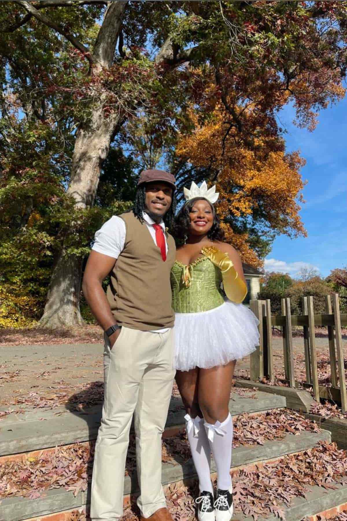 Two people in costumes stand outdoors on wooden steps with autumn trees in the background. One wears a vest and cap, the other a green bodice, white tutu, and a crown-like headpiece.