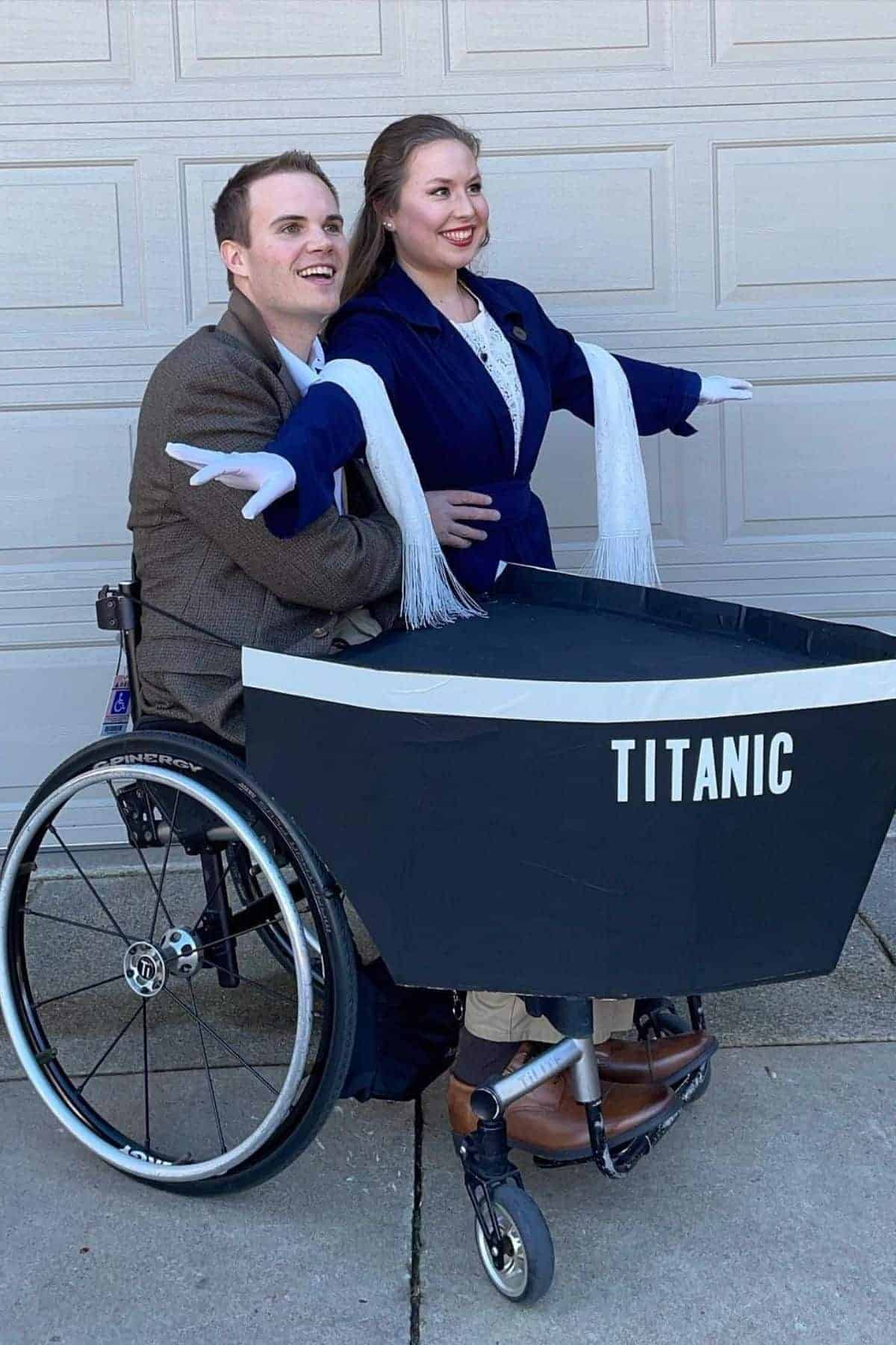 Two people recreate the Titanic movie pose in front of a garage, with one person in a wheelchair inside a cardboard boat labeled “Titanic.”.