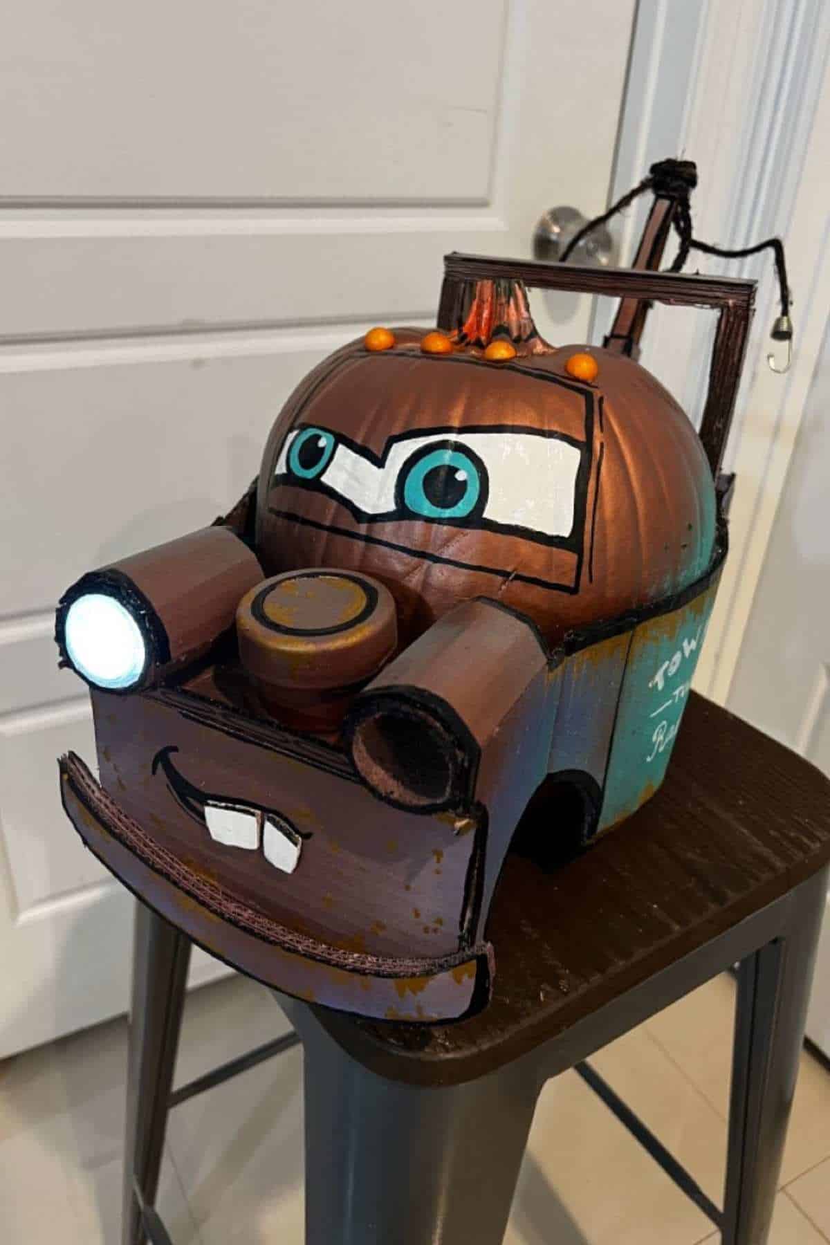 A pumpkin painted and crafted to look like a tow truck character with blue eyes, a smiling mouth, and headlights, placed on a stool indoors.