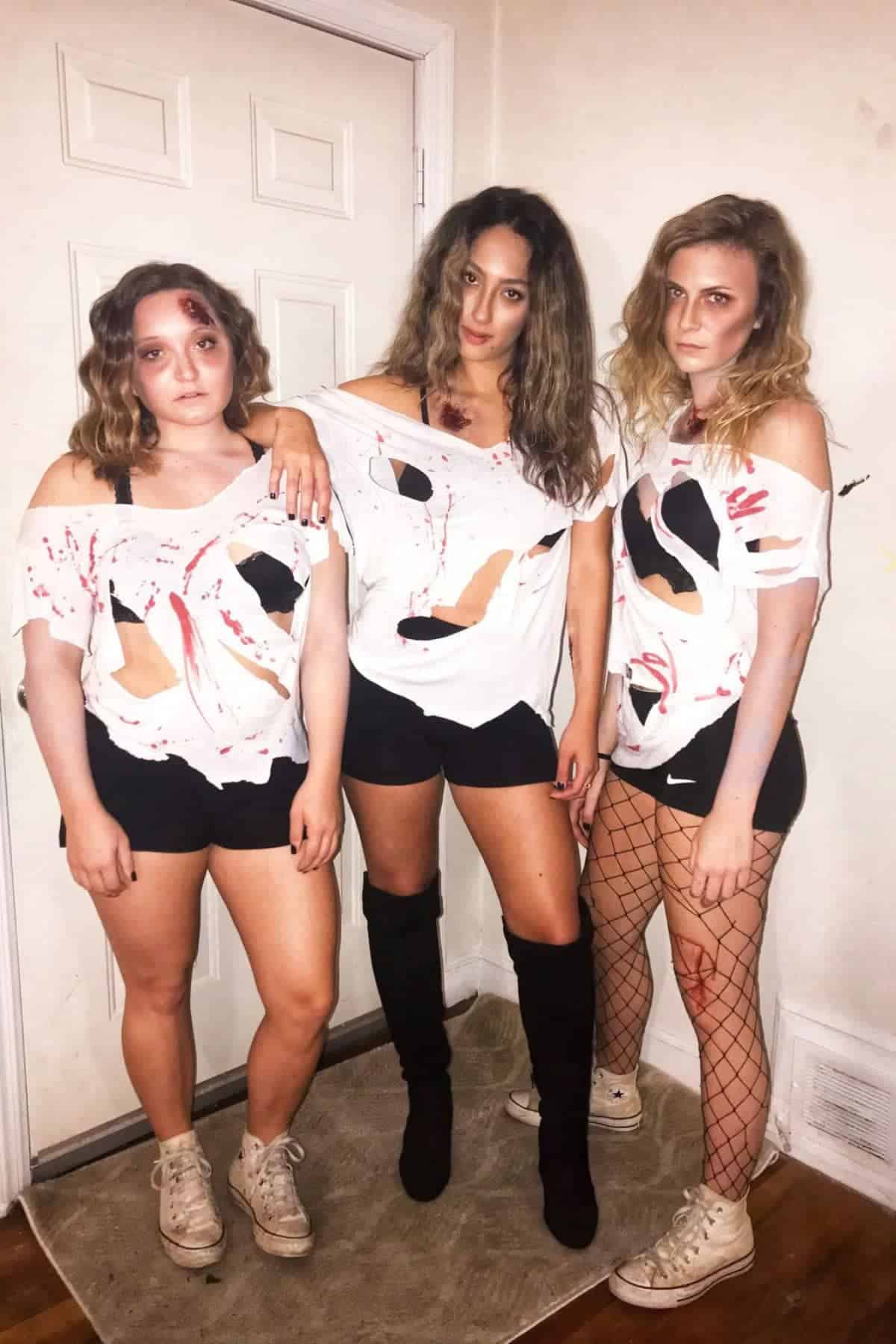 Three women stand indoors wearing torn white shirts with fake blood, black shorts, and distressed accessories, appearing as if dressed in zombie-themed or horror-inspired costumes.