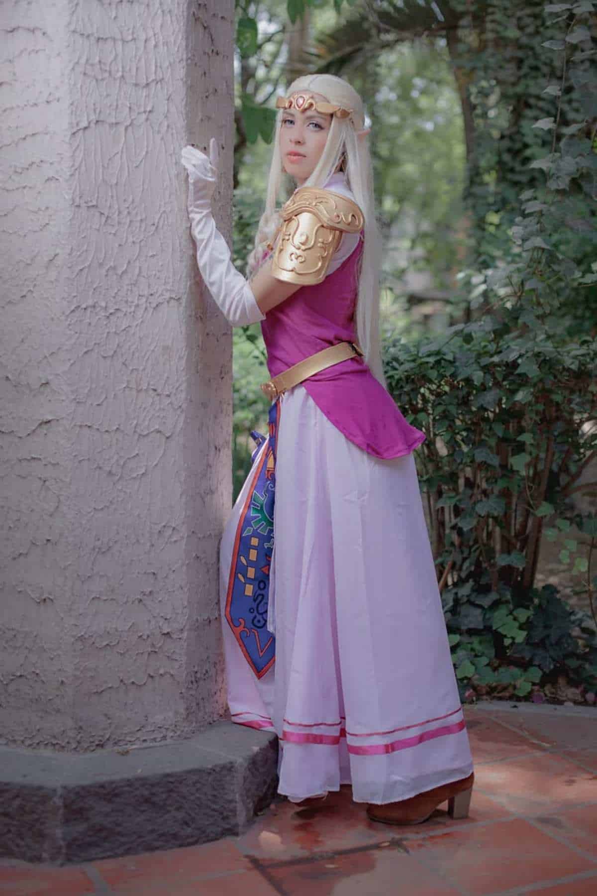 Person in a fantasy costume inspired by Zelda with long blonde hair, gold armor, pink top, and long white skirt stands by a textured column in a garden setting.