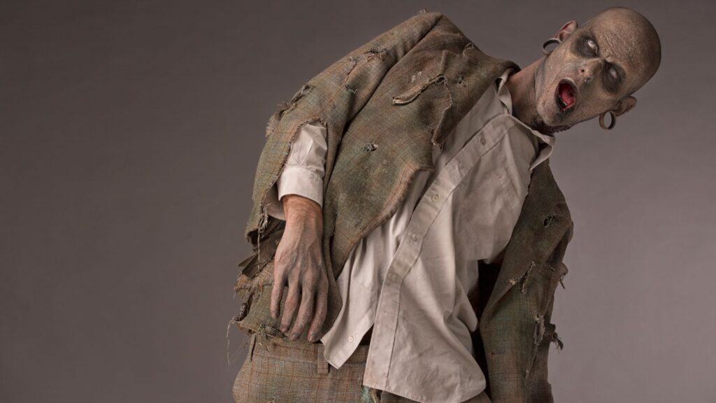 Person dressed as a zombie with tattered clothes and theatrical makeup, leaning to the side against a gray background.