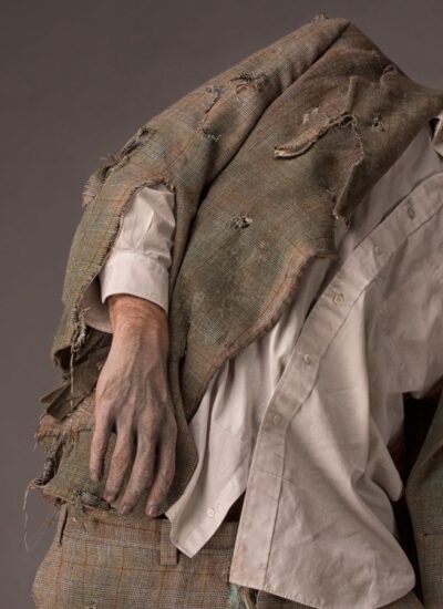 Person dressed as a zombie with tattered clothes and theatrical makeup, leaning to the side against a gray background.
