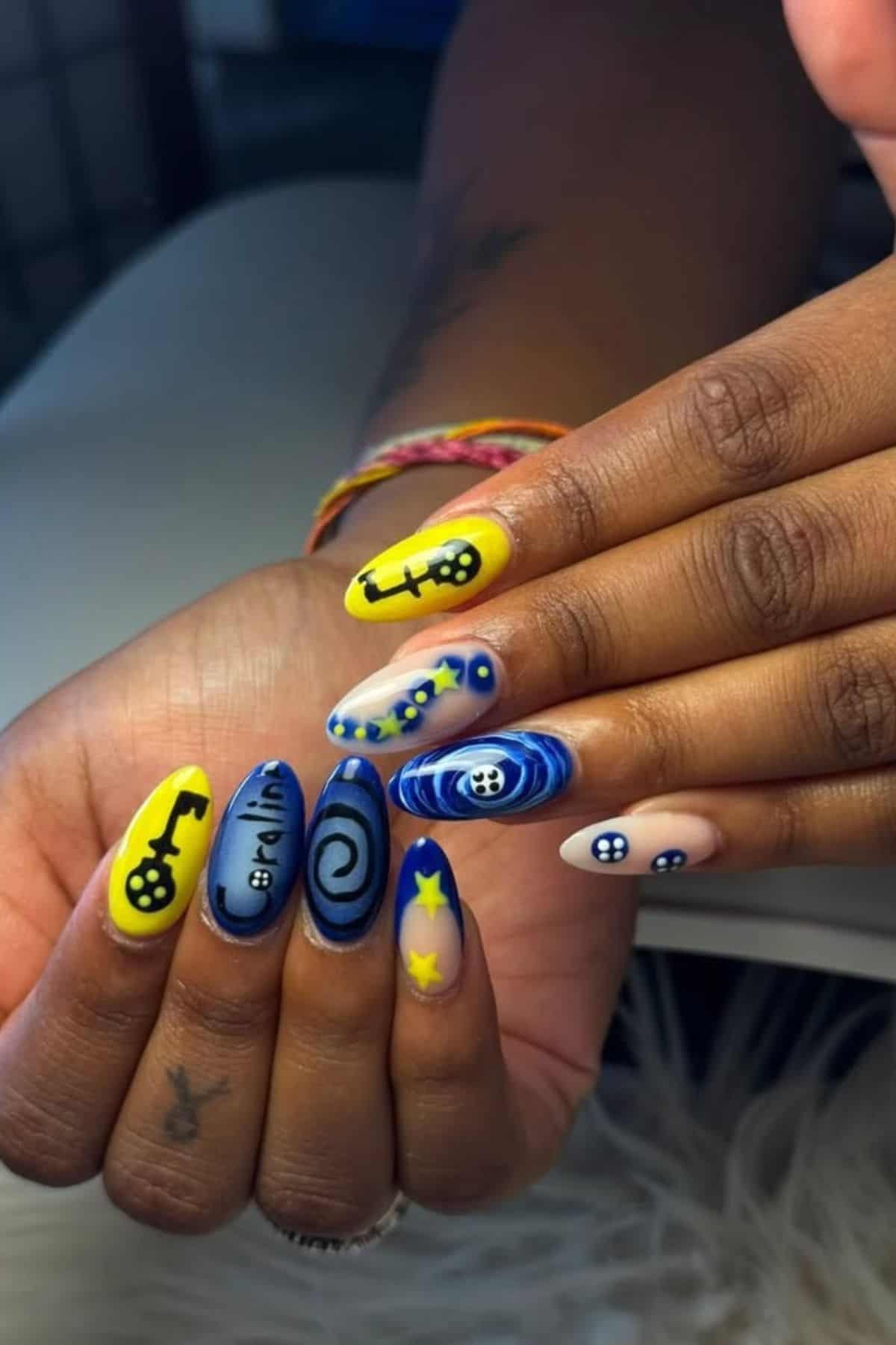 Hands with long, pointed nails painted in blue and yellow with key, eye, swirl, and star designs; background features part of a white table.