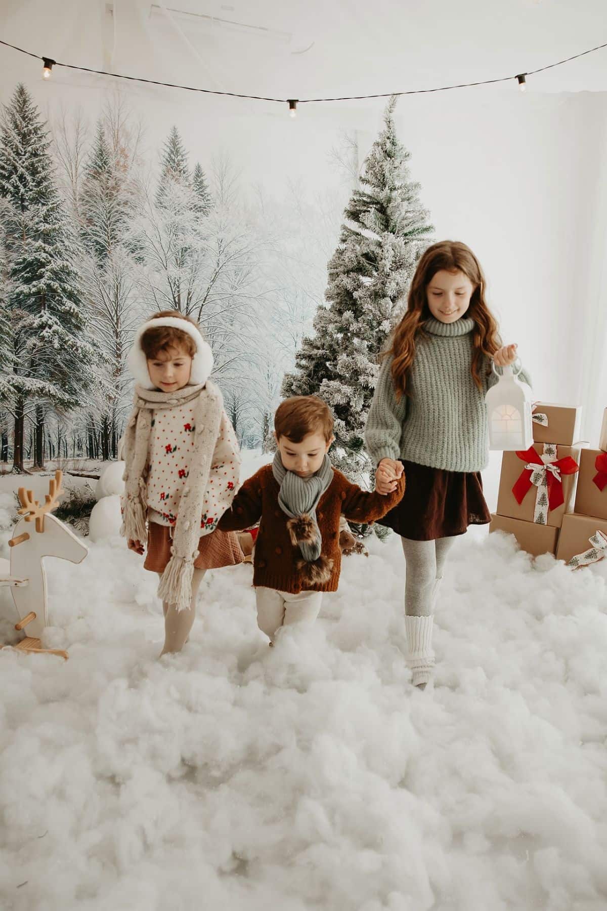 Three children wearing winter clothes walk hand-in-hand on artificial snow in a room decorated with Christmas trees and presents.