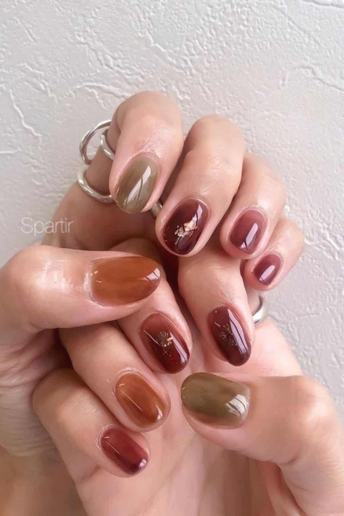 Hands with short, oval nails painted in earthy shades of brown, green, and mauve, some with minimal gold leaf accents. The person is wearing silver rings.