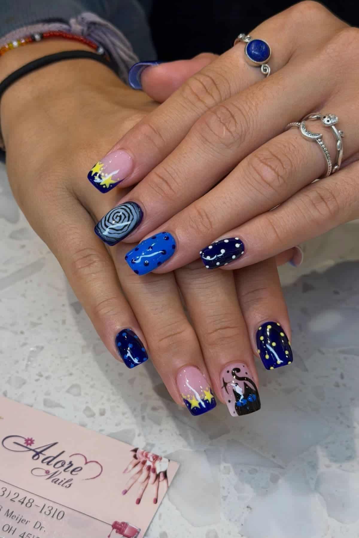 Hands with manicured nails featuring starry night and Van Gogh-inspired designs in blue, yellow, and black. Silver rings and a blue stone ring are worn on the fingers. A business card is visible.