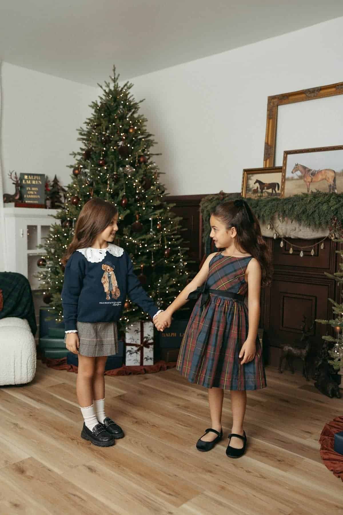 Two young girls holding hands in a decorated living room with a Christmas tree, wearing plaid outfits and standing on a wooden floor.