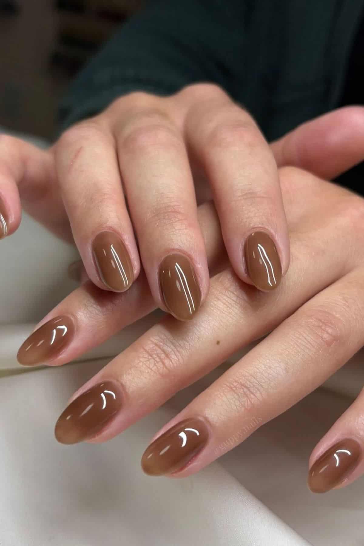 Close-up of hands with glossy, almond-shaped brown gel nail polish on natural nails, displayed against a neutral background.