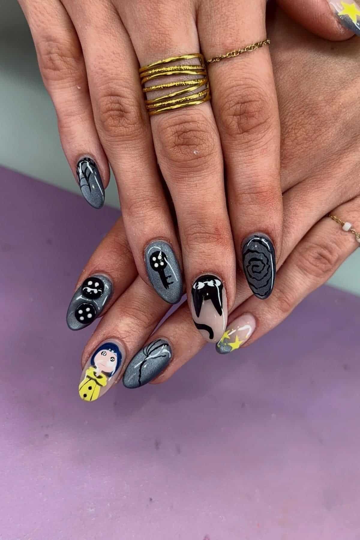 Hands with gray-themed nail art, including designs of a key, cat, face, swirl, and stars. Some nails feature black and white patterns and a yellow coat. Person wears gold and silver rings.