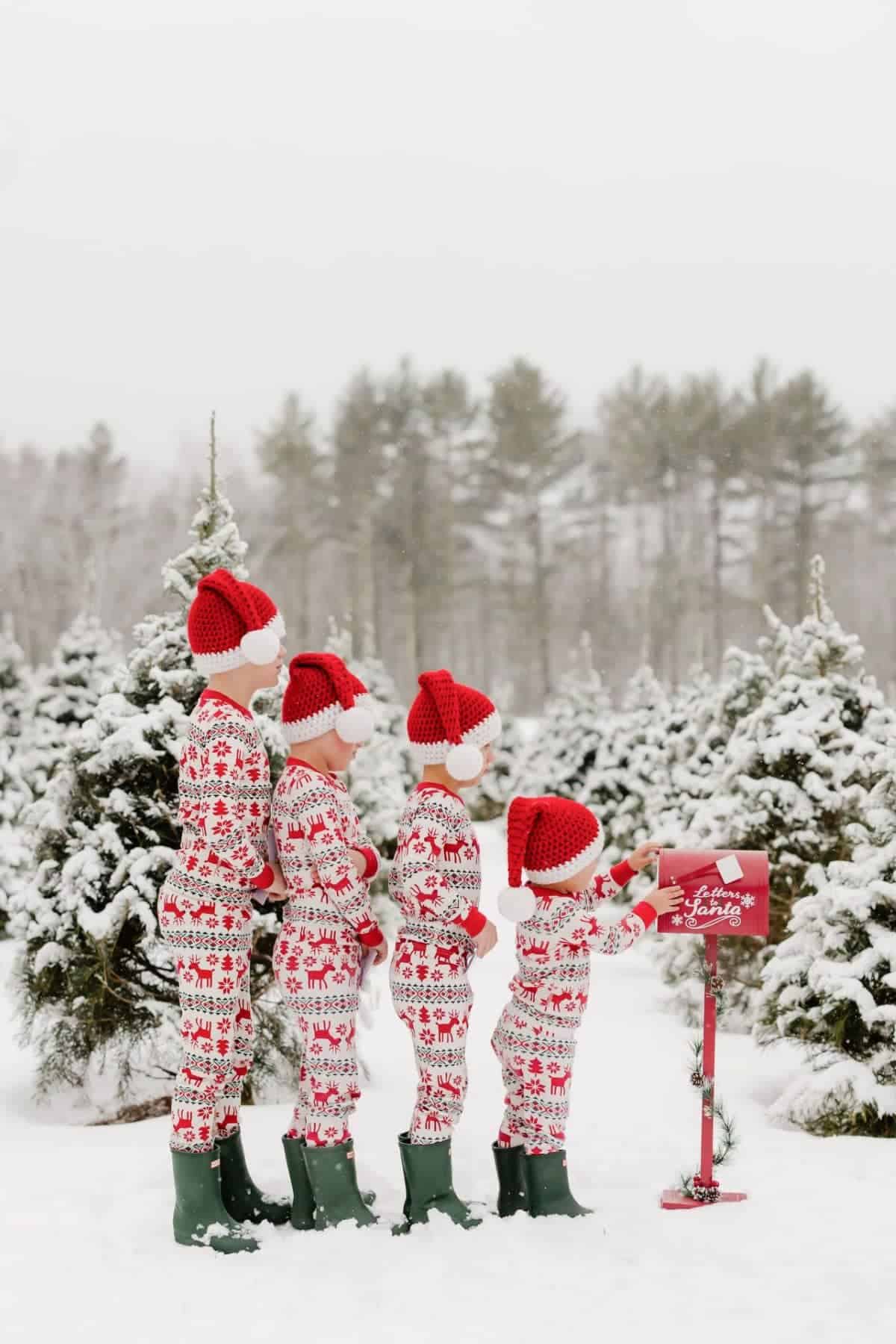 Four people in matching Christmas pajamas and Santa hats stand in a snowy tree field as a child places a letter into a red mailbox labeled “Letters to Santa.”.