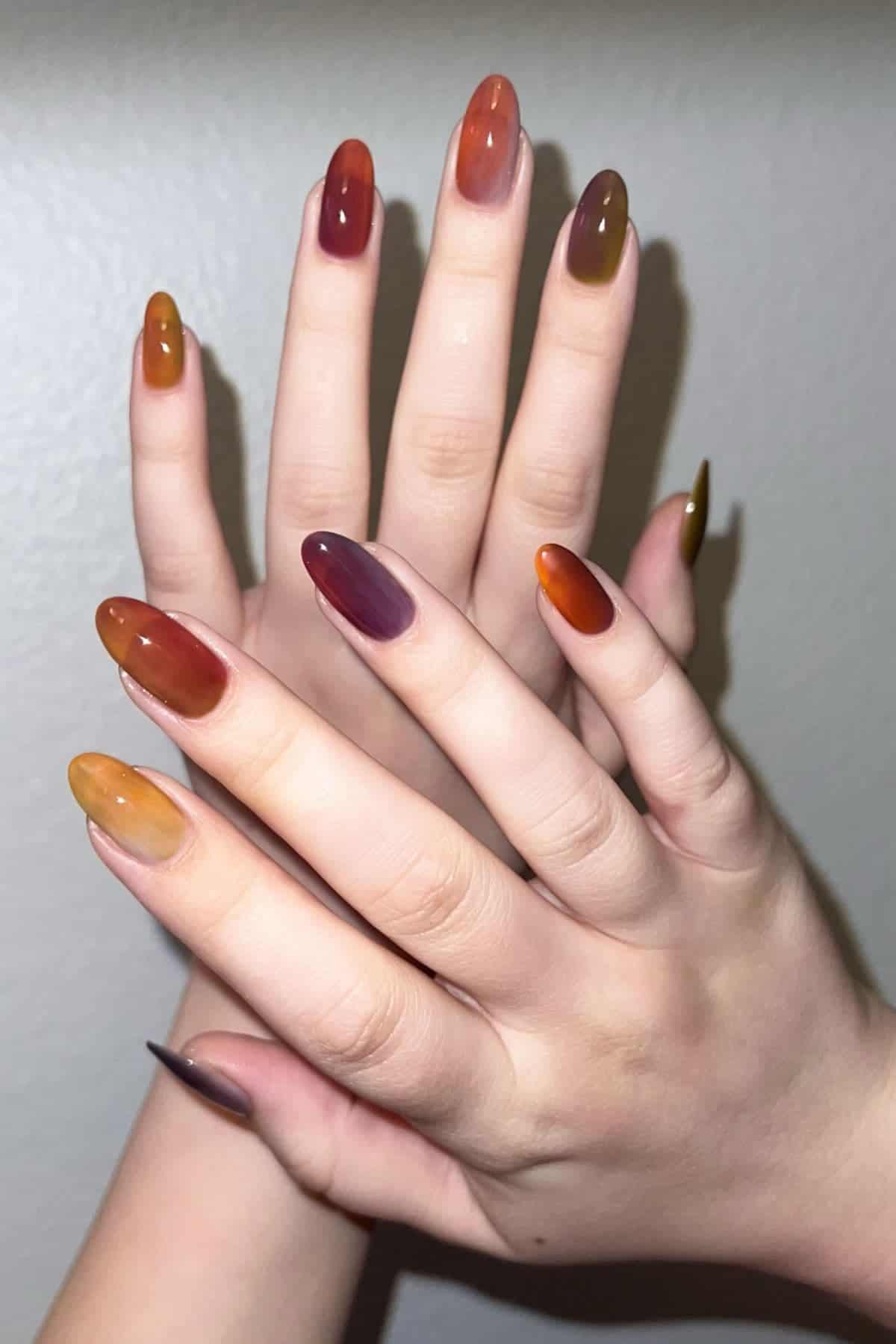 Two hands with long, almond-shaped nails painted in a gradient of autumn colors including red, orange, yellow, and olive green, displayed against a plain light background.