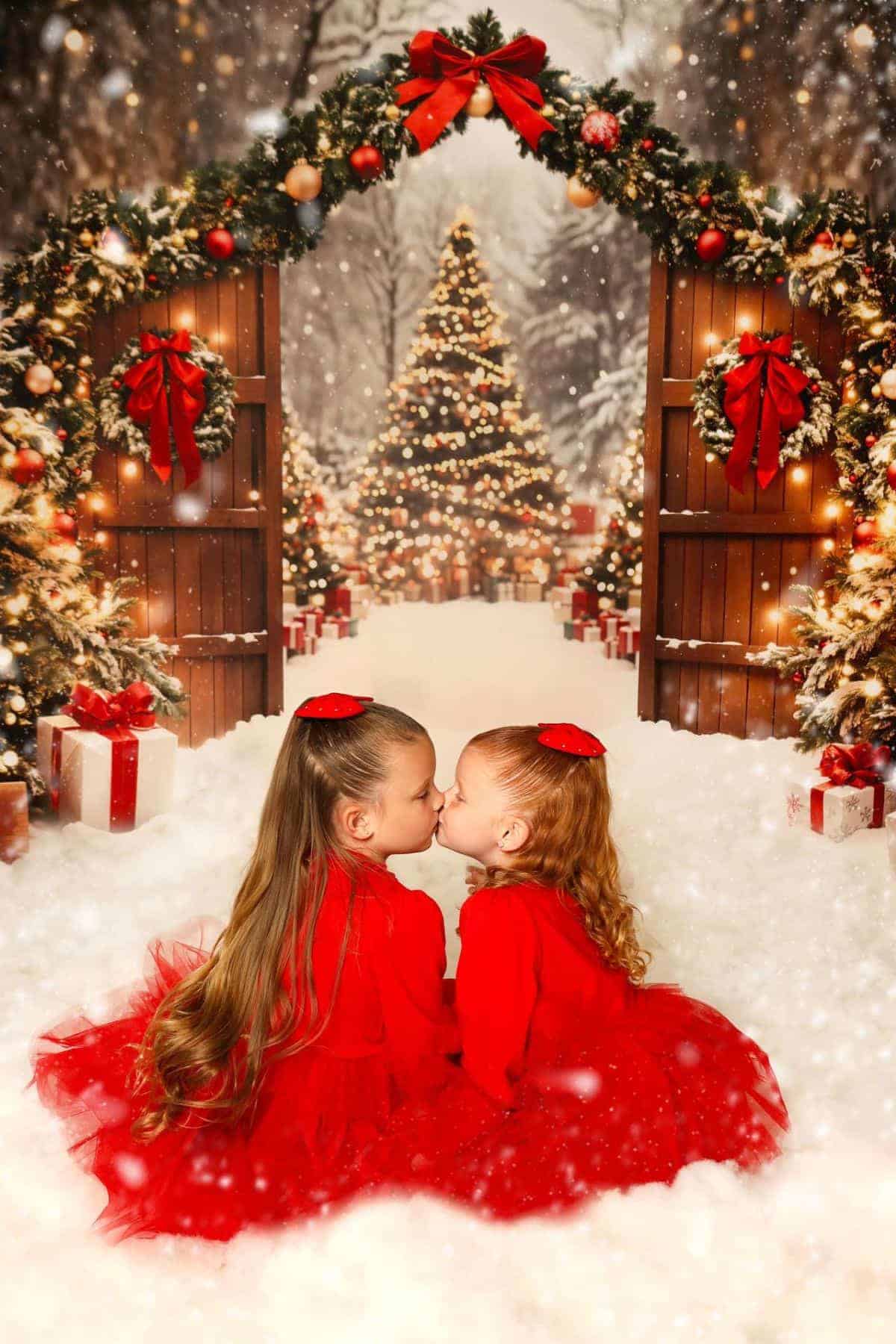 Two young girls in red dresses sit in snow, touching noses, with decorated Christmas trees, gifts, and festive arches behind them in a snowy outdoor setting.
