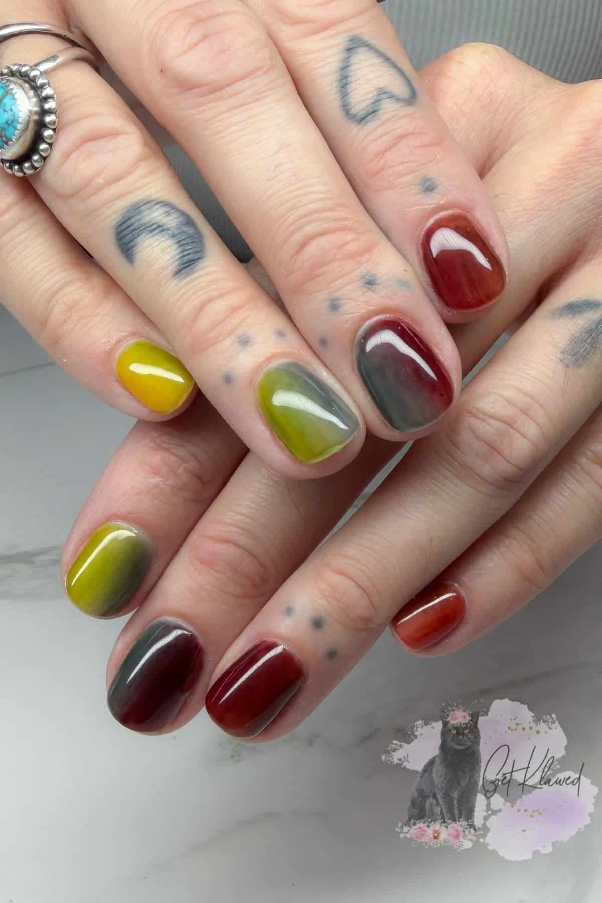 Hands with olive green, burgundy, and yellow gradient nails, visible hand tattoos including hearts and dots, and a silver ring with a turquoise stone.