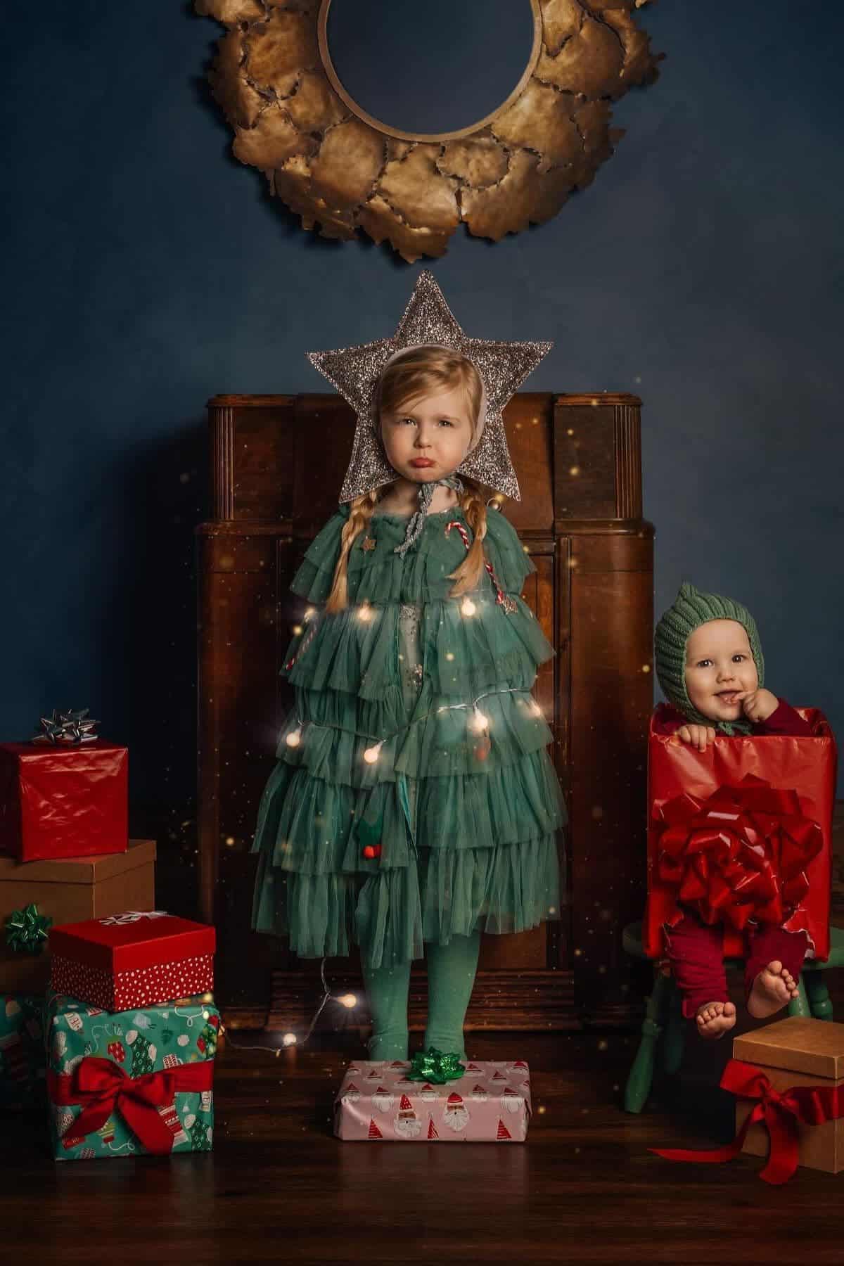 A young girl dressed as a Christmas tree stands on a present, with string lights and a star hat, next to a baby in a gift box costume, surrounded by wrapped presents.