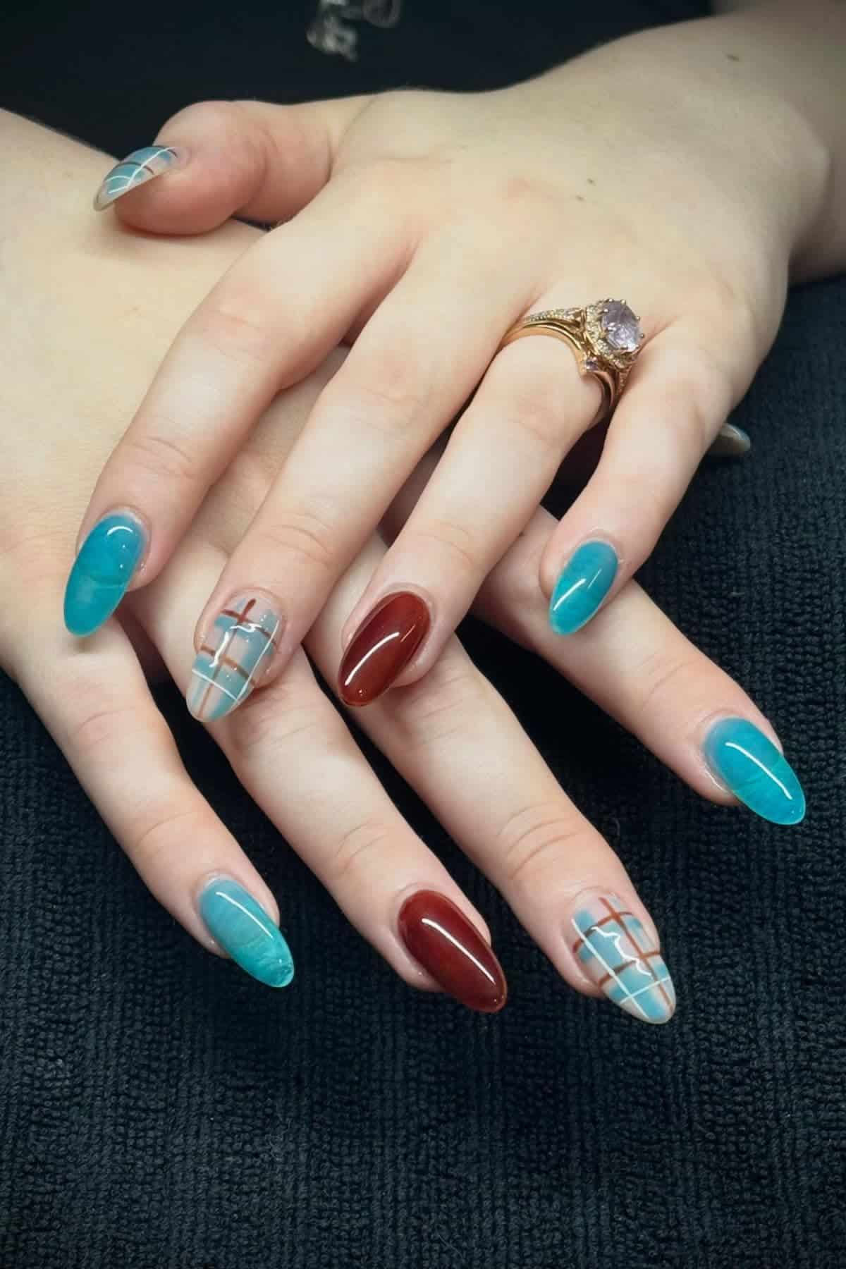 Hands with long, almond-shaped nails painted in teal, brown, and plaid designs on a dark surface. A gold ring with a purple stone is on one finger.