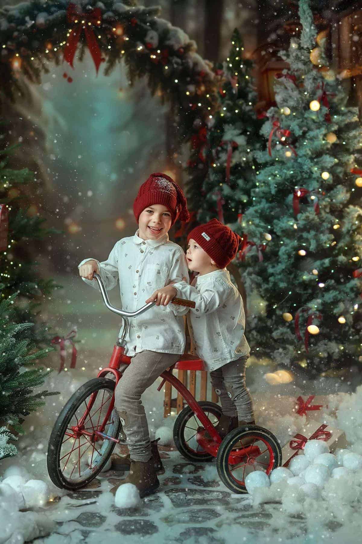 Two young children in matching white shirts and red hats stand with a tricycle among Christmas trees and festive decorations in a snowy, decorated setting.