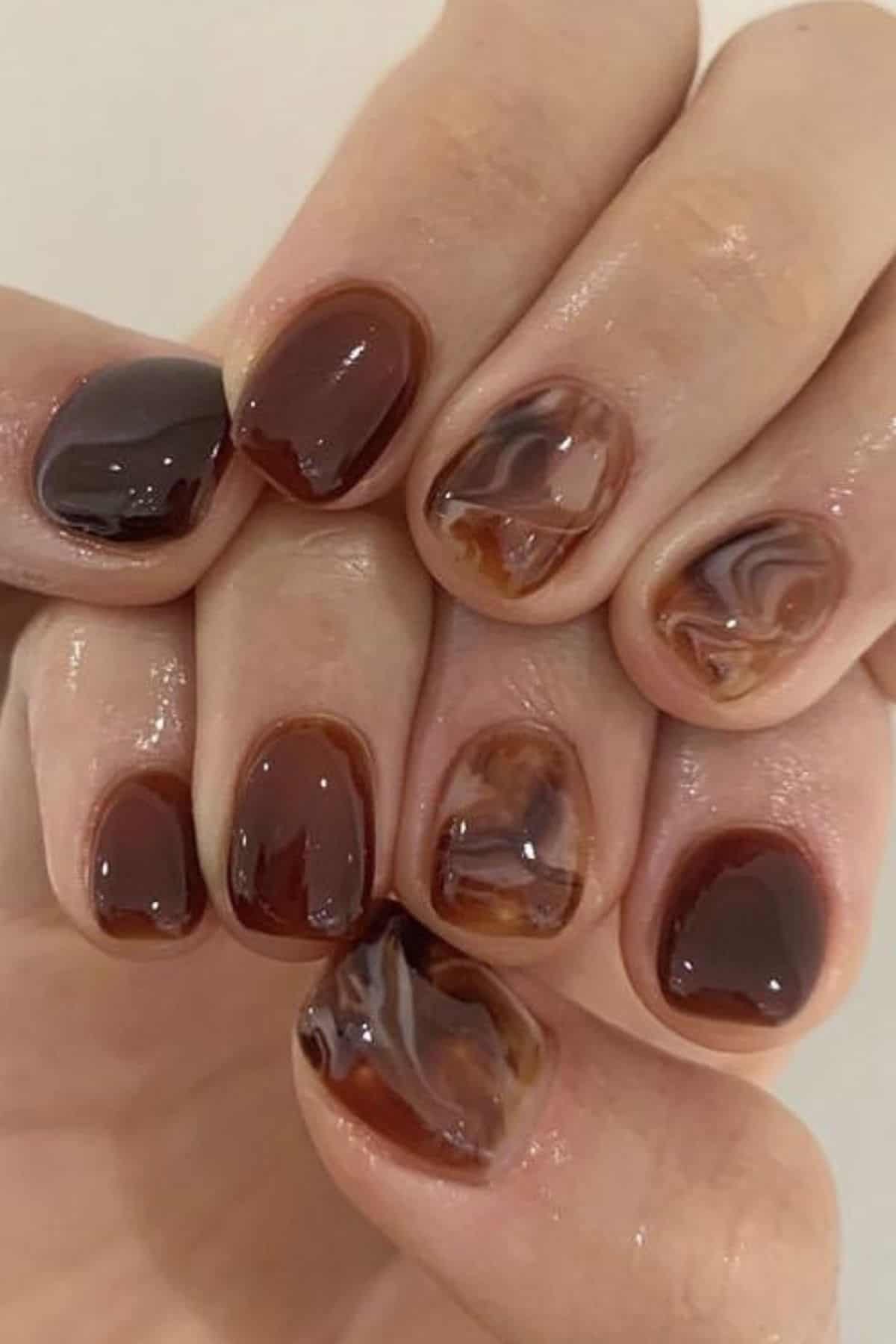 Close-up of hands with short, glossy brown and marbled brown nail art, featuring a mix of solid and swirled patterns on each fingernail.