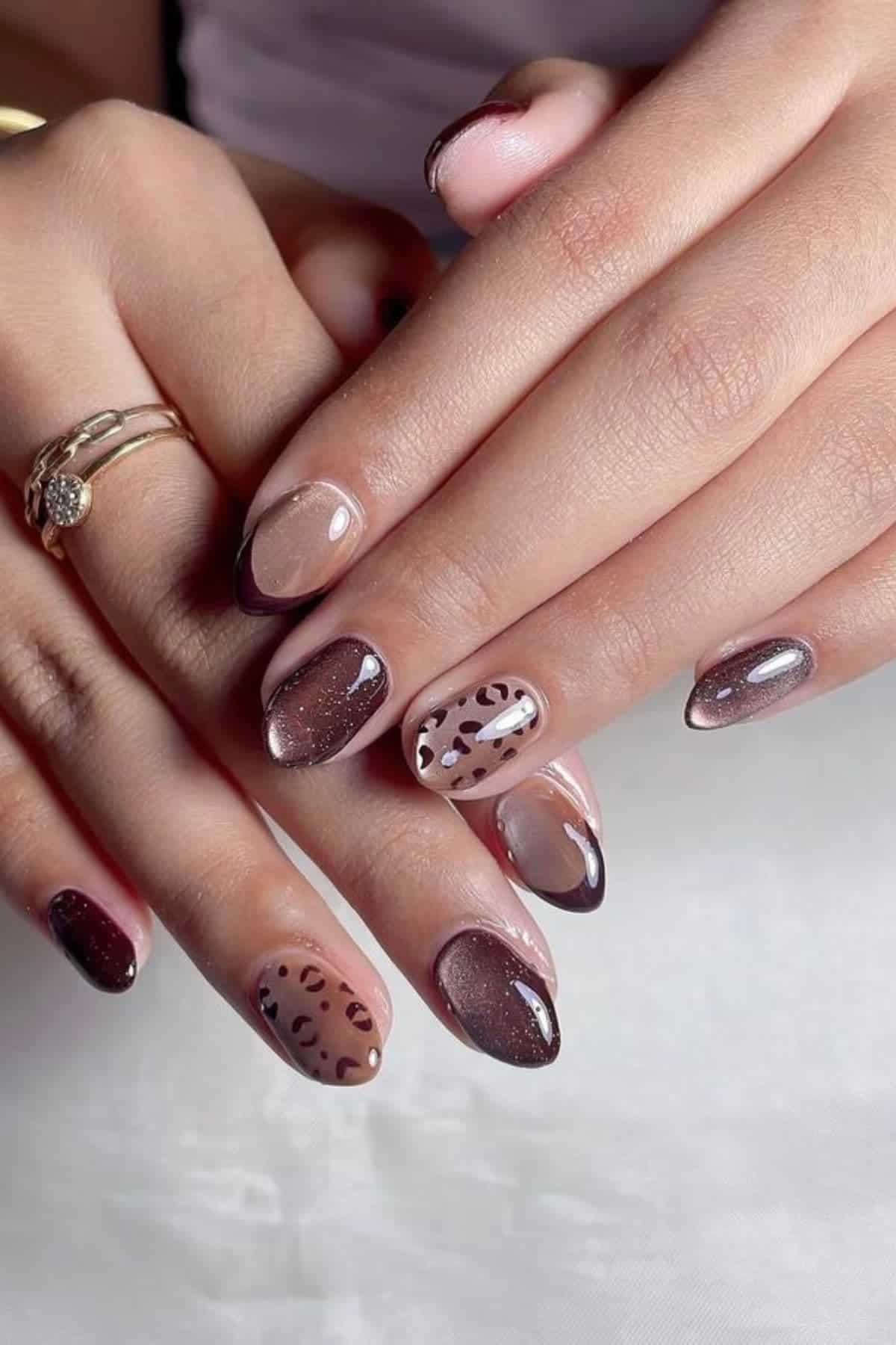 Hands with almond-shaped nails featuring a mix of brown, nude, glitter, and animal print designs. The person is wearing gold rings on the ring finger.