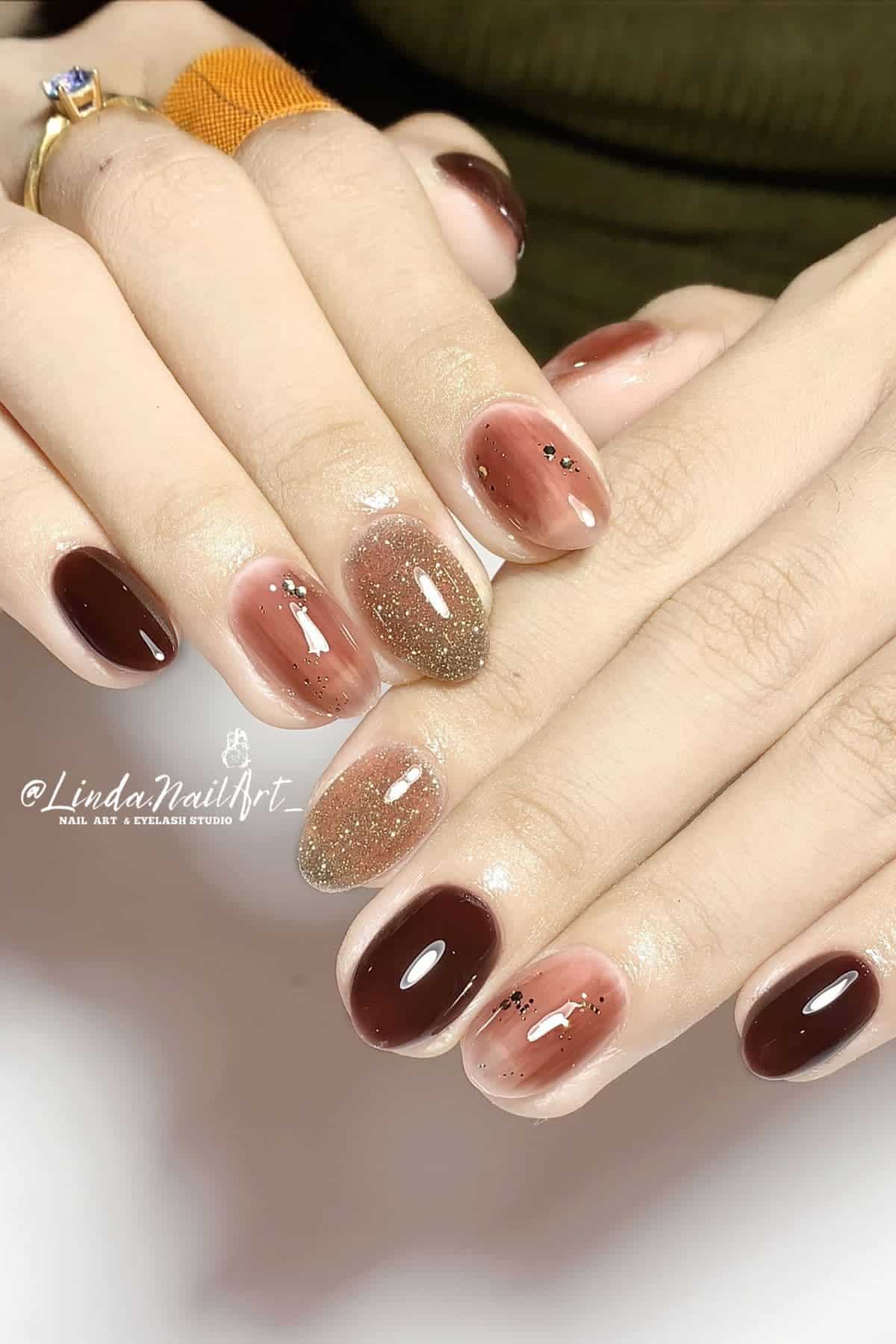 Hands with almond-shaped nails featuring dark brown, nude, and glittery polish, some with small black and gold accents.