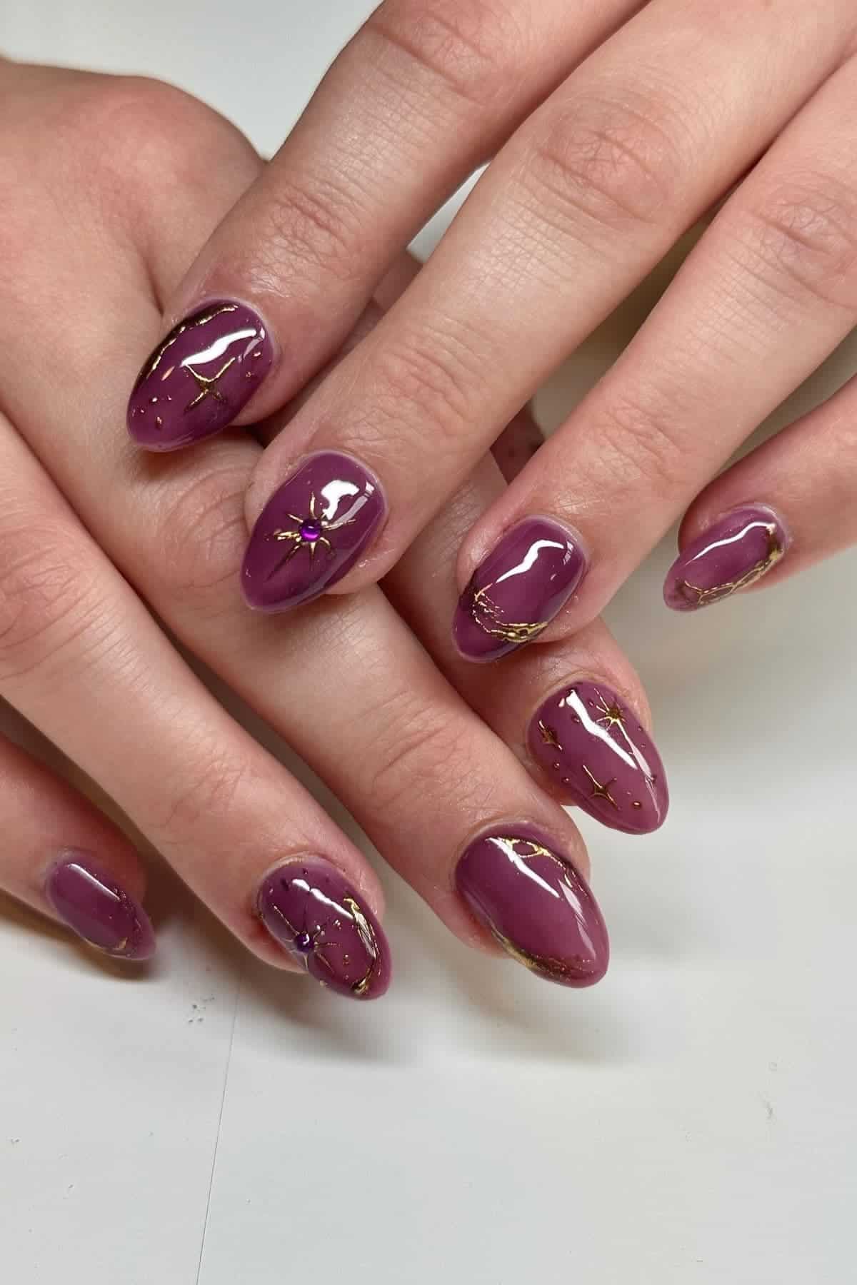 Hands with almond-shaped nails painted glossy mauve, accented with gold foil and intricate line designs.