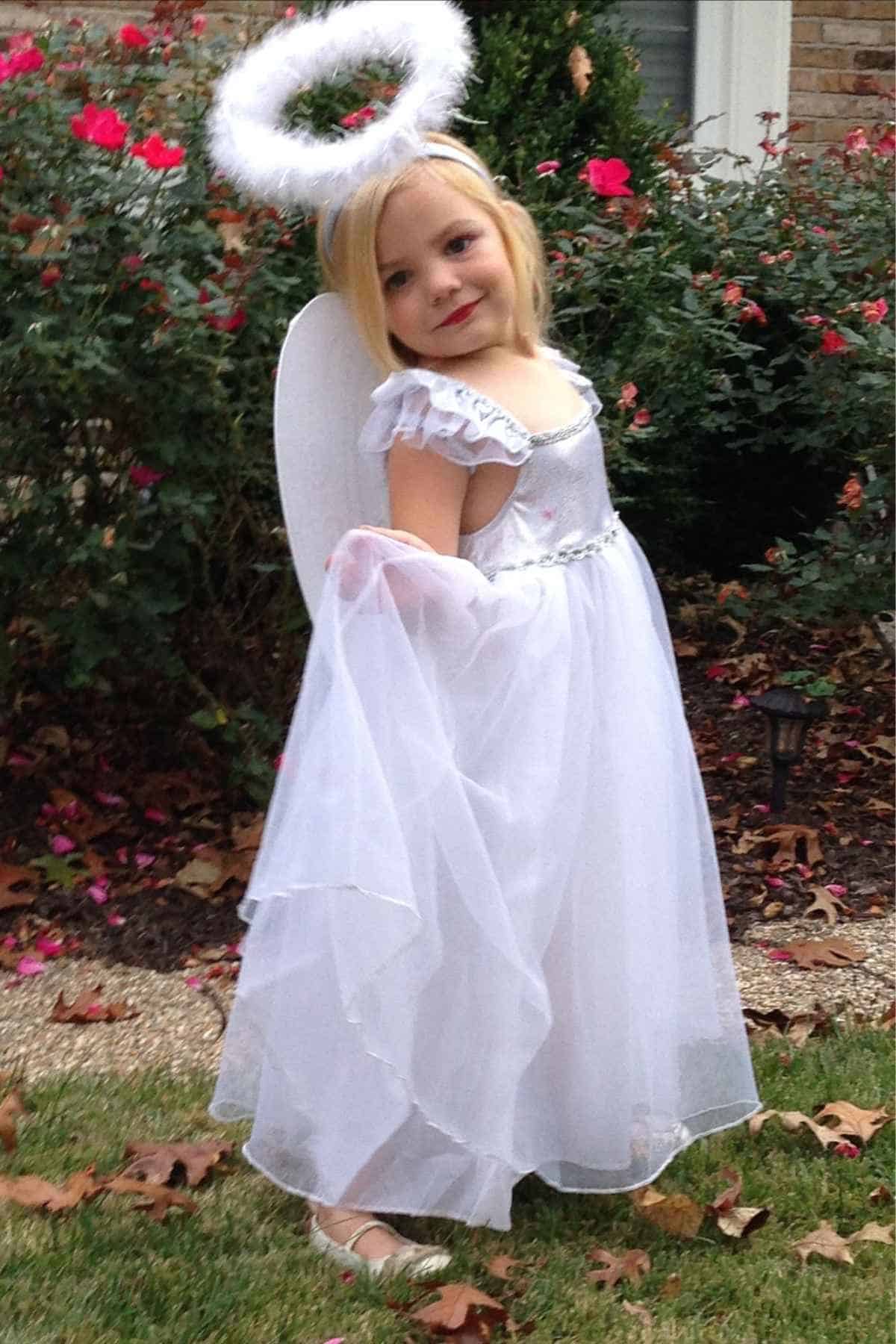 A young child in a white angel costume with wings and a halo poses outside in a garden with red flowers and green bushes.