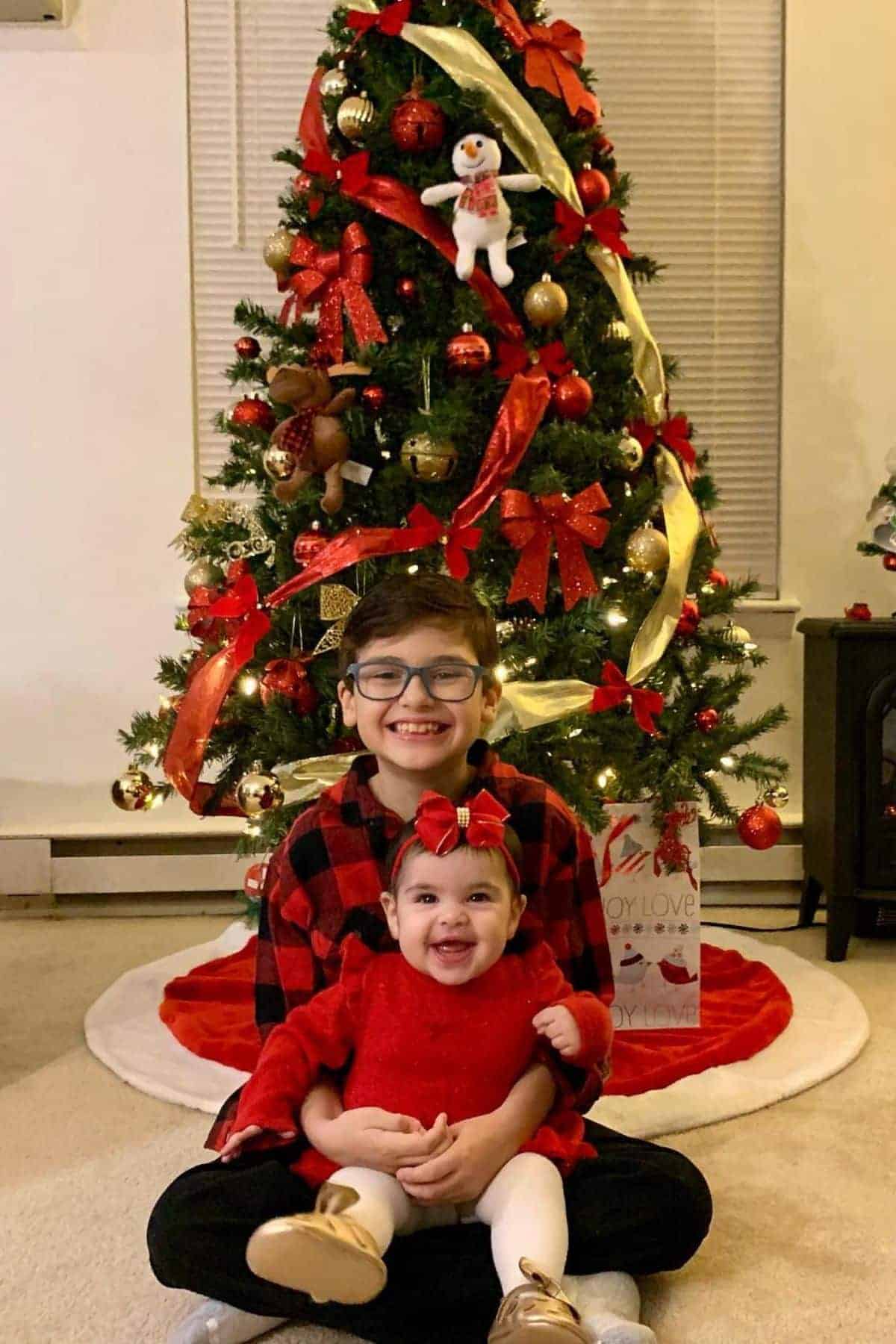 Two children in red outfits sit smiling in front of a decorated Christmas tree with ornaments, ribbons, and gifts underneath.