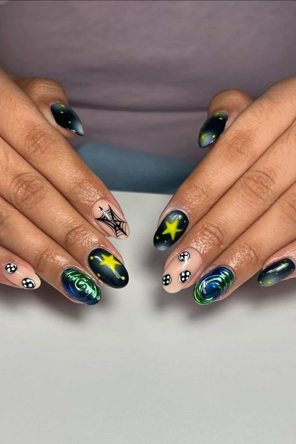 Hands with manicured nails featuring designs of stars, spider webs, swirls, and dots in black, green, yellow, and blue tones on a neutral background.
