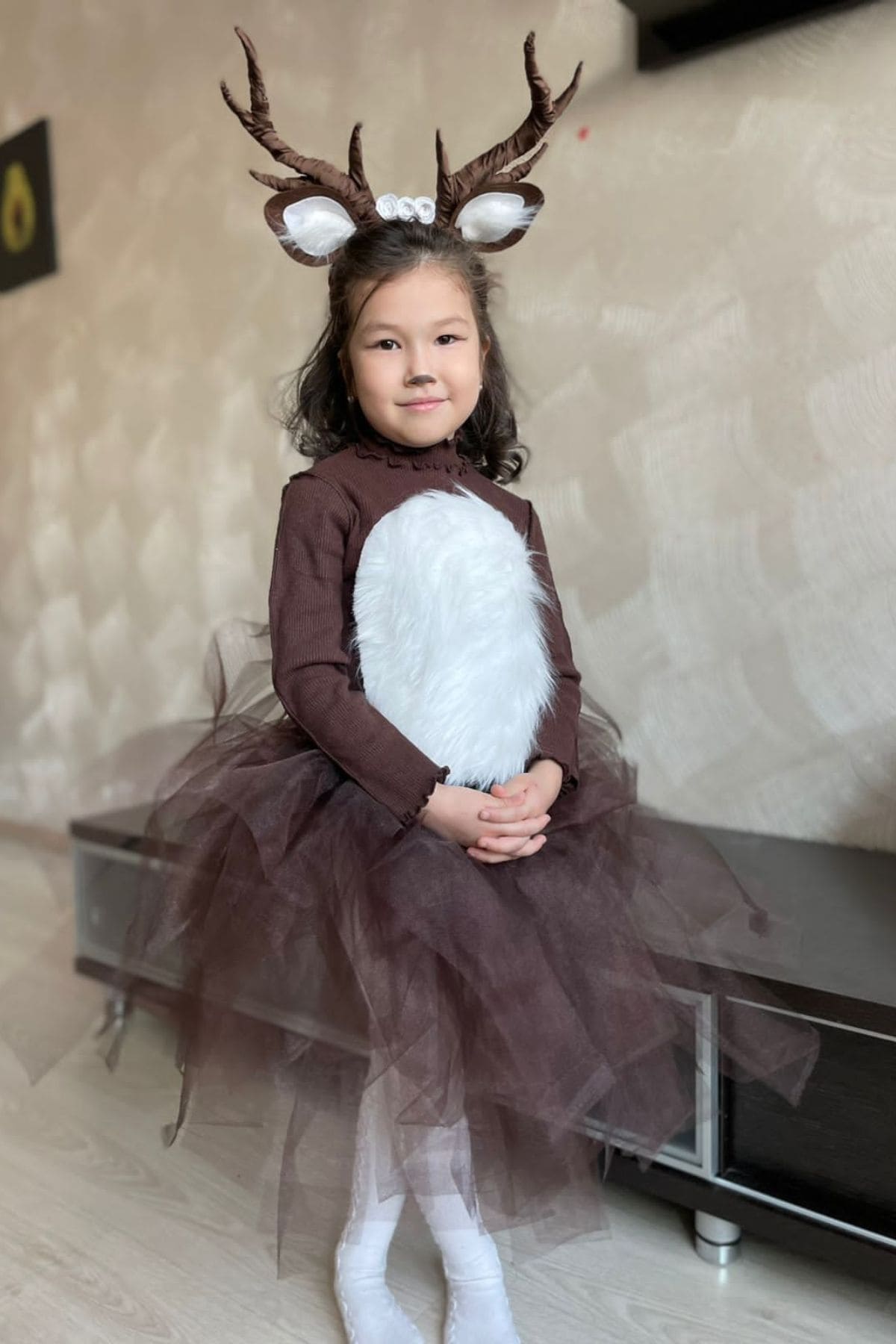 A young girl in a deer costume with antlers, a brown dress, and a white furry patch on the front, sits on a bench with her hands folded in her lap.