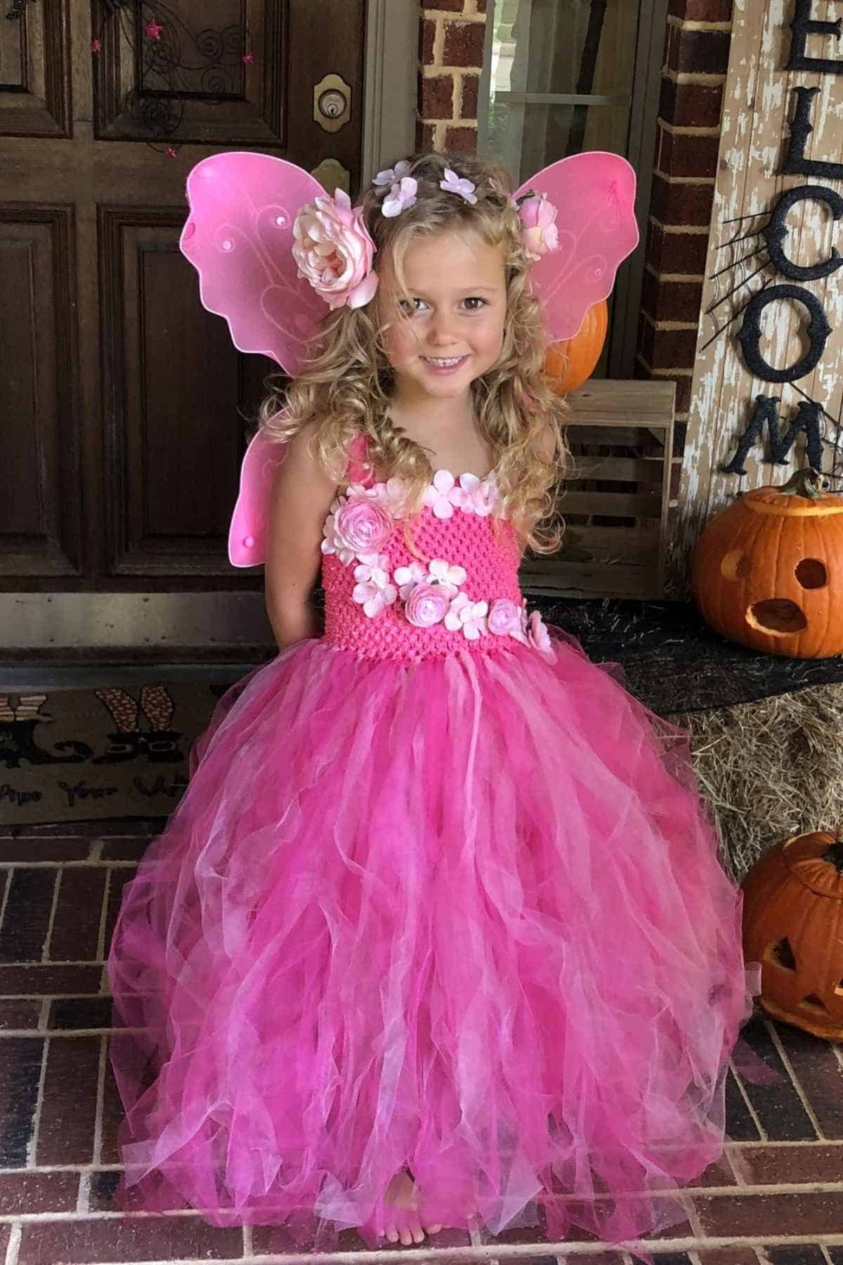 Young girl in a pink fairy costume with wings, flower headband, and tulle dress stands barefoot on a porch decorated with carved pumpkins.