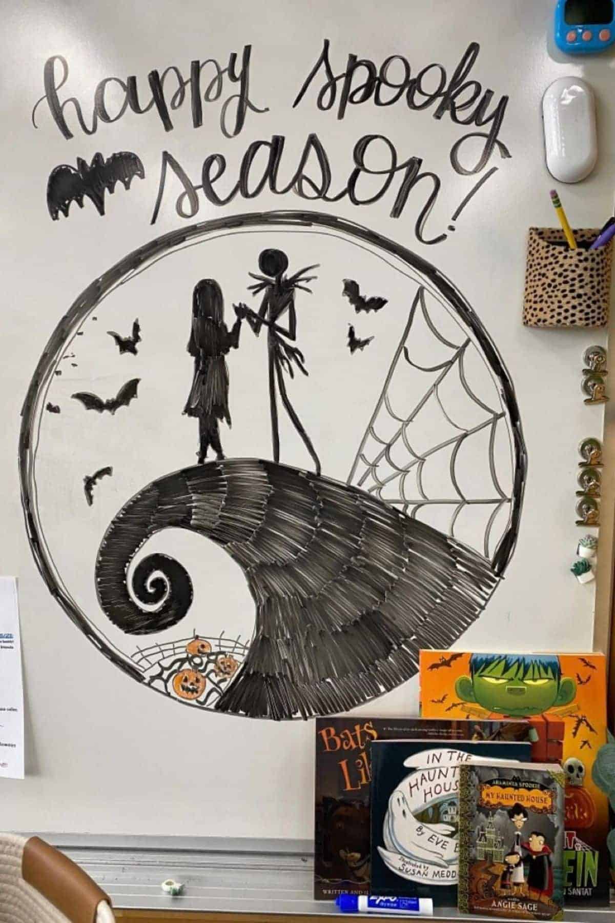 Whiteboard with “happy spooky season!” and a drawing of two Halloween characters on a spiral hill, bats, spiderweb, and pumpkin. Halloween books are displayed below the drawing.