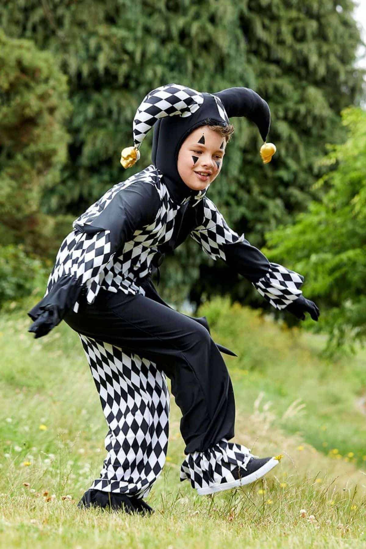A child in a black and white diamond-patterned jester costume crouches outdoors on grass, with trees in the background.