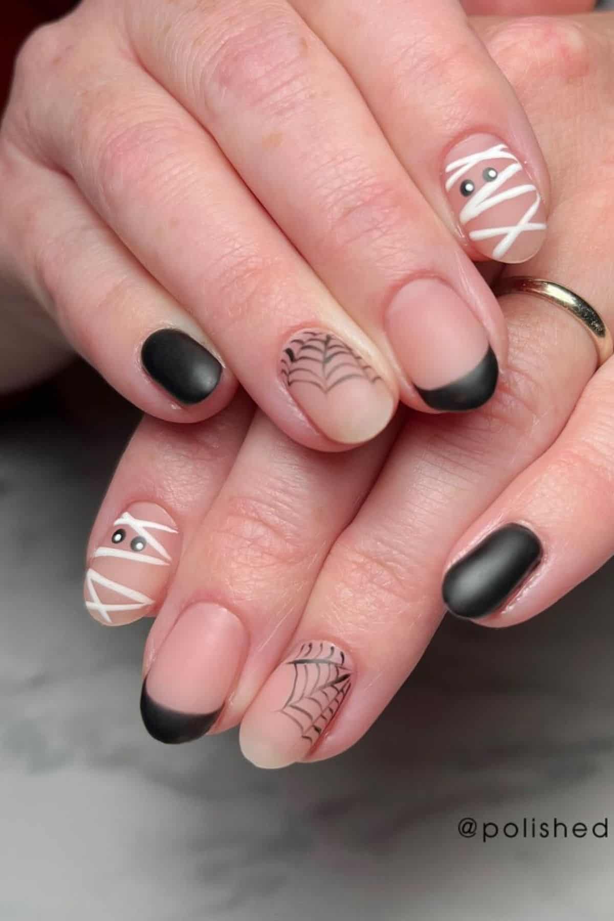 Hands with Halloween-themed nail art featuring black-tipped nails, spiderweb designs, and mummy faces on a matte nude base.
