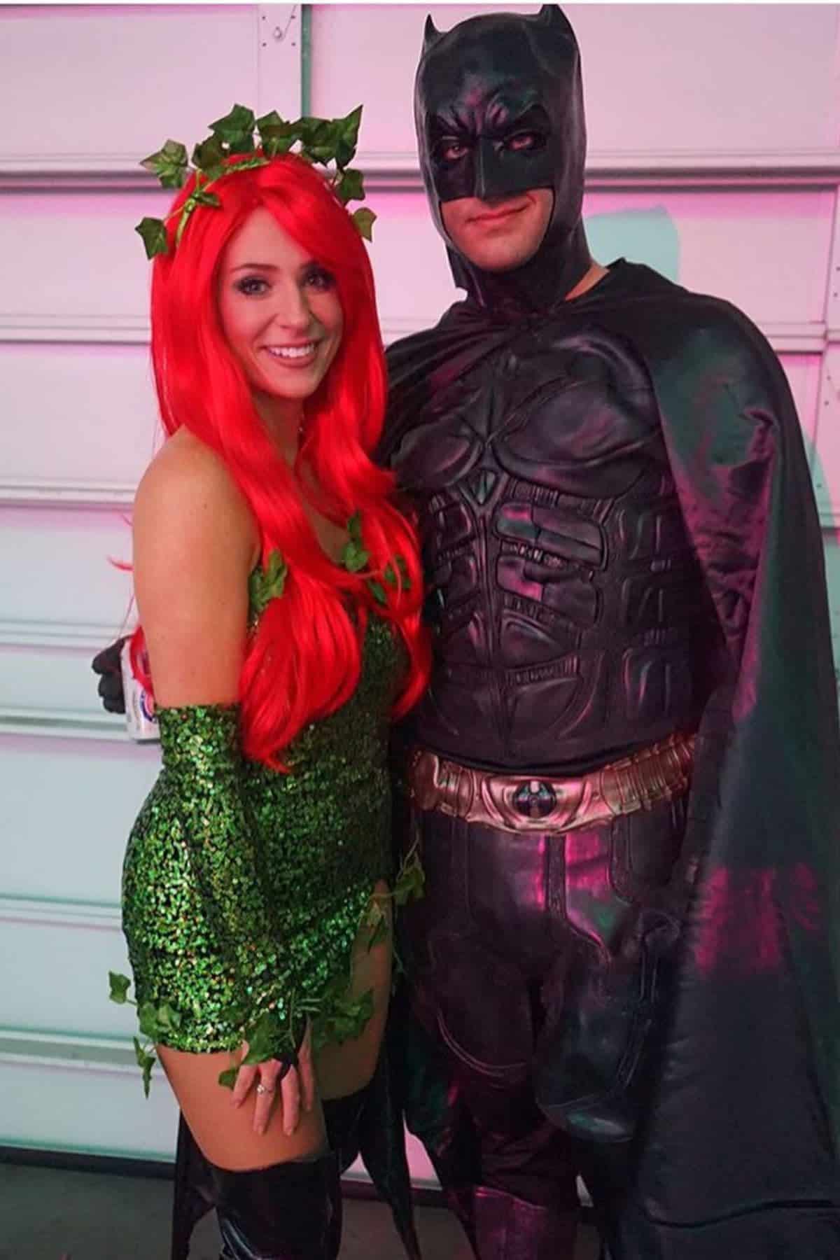 Two people in costumes pose together. One is dressed as Poison Ivy with a red wig and green outfit, and the other as Batman in a black suit and cape. They are standing indoors in front of a garage door.