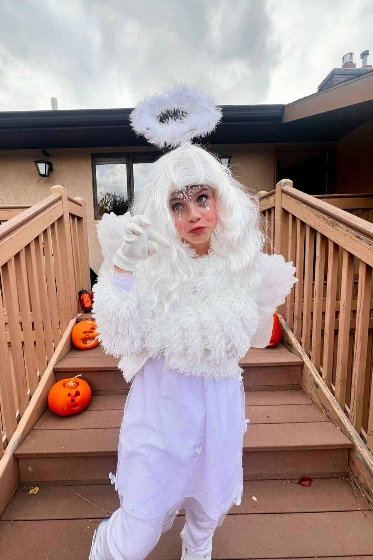 Person in a white angel costume with wings and halo stands on outdoor steps decorated with carved pumpkins.